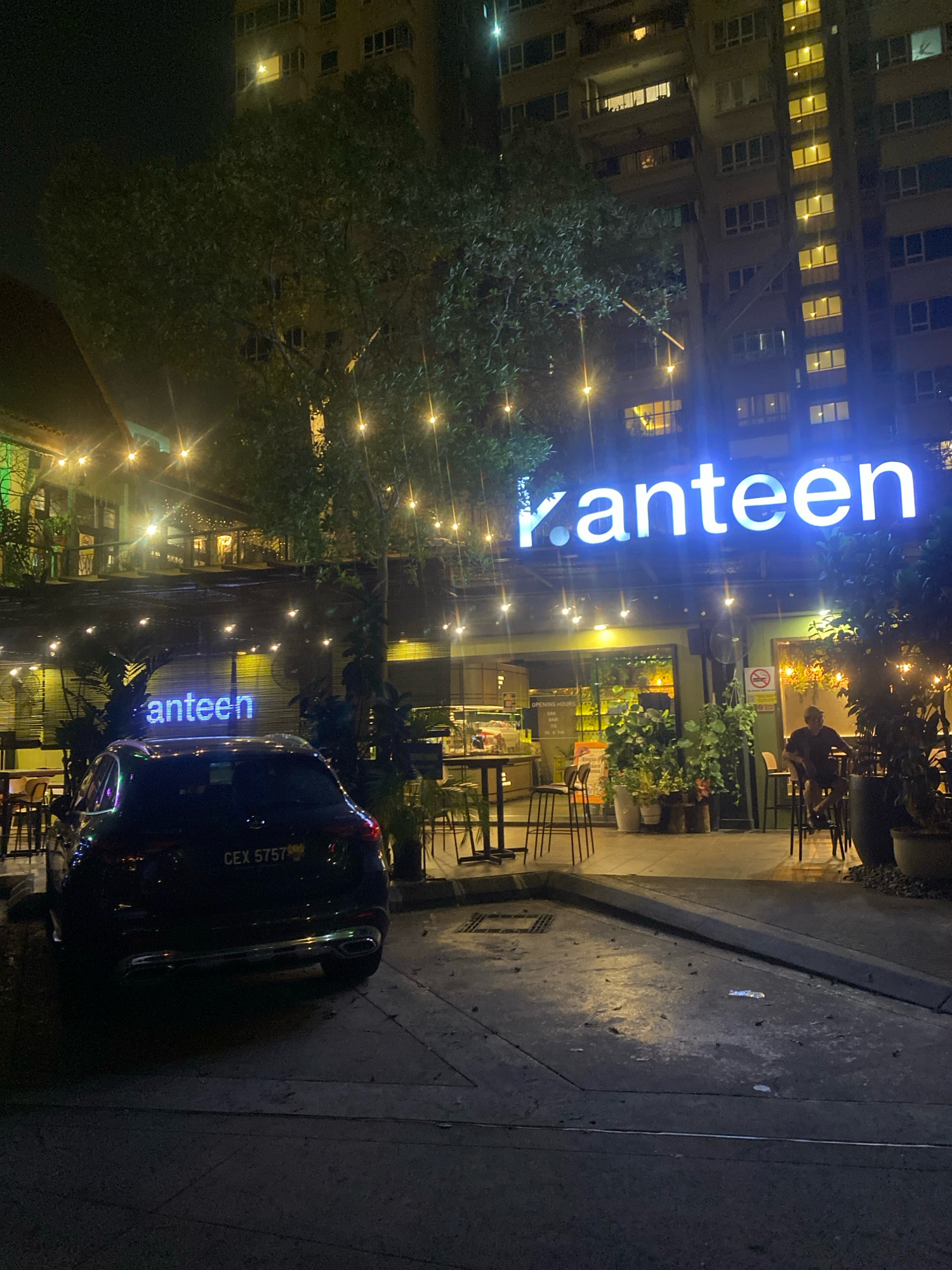At night in Kuala Lumpur, Malaysia, a black Mercedes-Benz GLE SUV is parked in front of a restaurant called "Kanteen." The restaurant features a neon sign with the name "Kanteen" illuminated in blue. The building behind the restaurant is a tall residential apartment complex with many windows, some of which are lit. The scene is illuminated by the neon sign, string lights, and the lights from the apartment building. A person is seated at a table outside the restaurant. The ground is paved and appears wet in some areas, possibly from recent rain or cleaning. The overall atmosphere is that of a modern urban setting at night, with a focus on the restaurant and its signage.