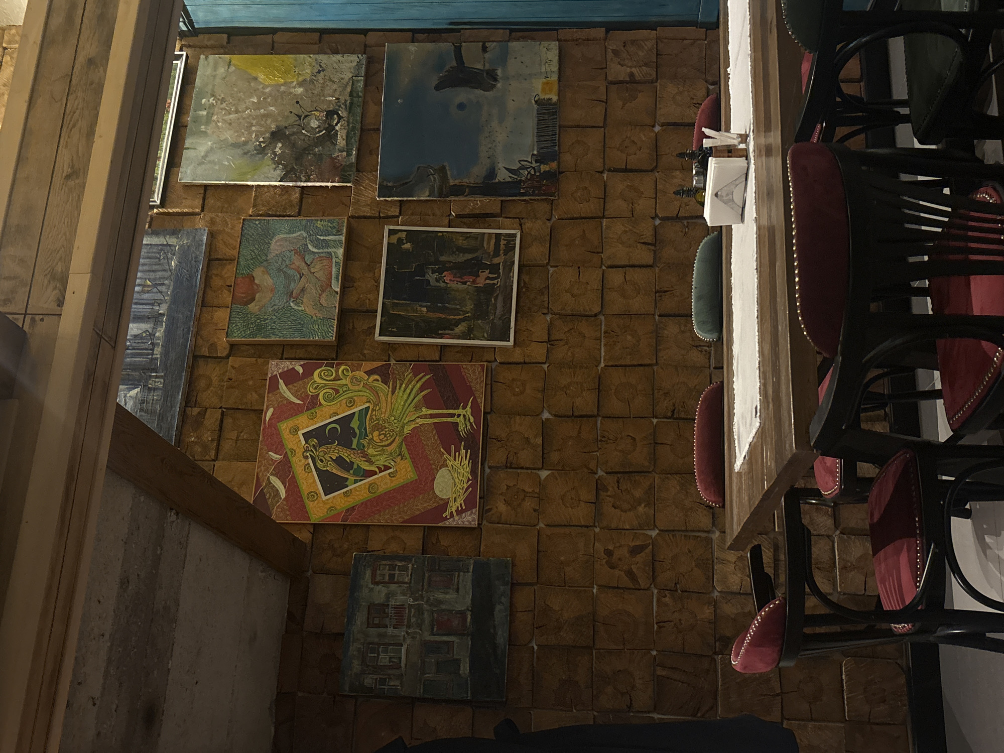 The image captures a scene inside a restaurant or cafe in Varna, Bulgaria. The walls are decorated with a collection of framed abstract paintings and drawings, mounted on a background of textured wood blocks. The space is lit with warm lighting. A dark wood table is visible along the right side of the image, with black metal framed chairs, featuring red and green cushioned seats. A table setting is present, including a napkin holder and cutlery. The perspective is from an elevated angle, looking down on the table and wall decorations.