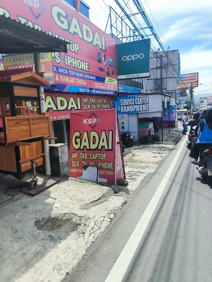 The image shows a street scene in Ciwidey, Indonesia, at the intersection of a main road and a side street. A large red sign with the word "Gadai" (pawn shop) in white letters is prominently displayed on the corner of a building. The sign is advertising pawn services for a variety of electronics, including HP, Tab, Laptop, SLR, TV, PS, and iPhone. There are also a few other signs, including one advertising a "Service Center Handphone". There are a few people walking down the street, including a woman in a blue shirt and a man wearing a black shirt. Several motorcycles are parked on the side of the road. The road surface is cracked and uneven, indicating a lack of maintenance. There are power lines overhead, and the sky is partly cloudy. A white painted line runs down the center of the street, separating the two lanes of traffic. The scene appears to be from a busy commercial area with a fair amount of foot traffic.