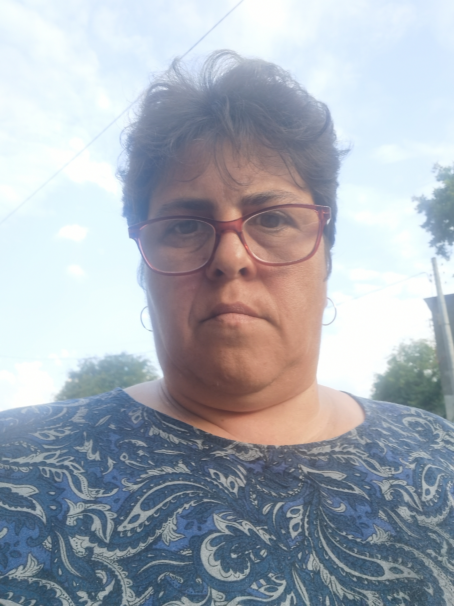 A woman wearing glasses and a blue paisley top stands outdoors, looking directly at the camera. The background shows a sky with clouds, trees, and a utility pole, suggesting an outdoor setting within the city of Lomets, Bulgaria. The lighting indicates it is daytime.