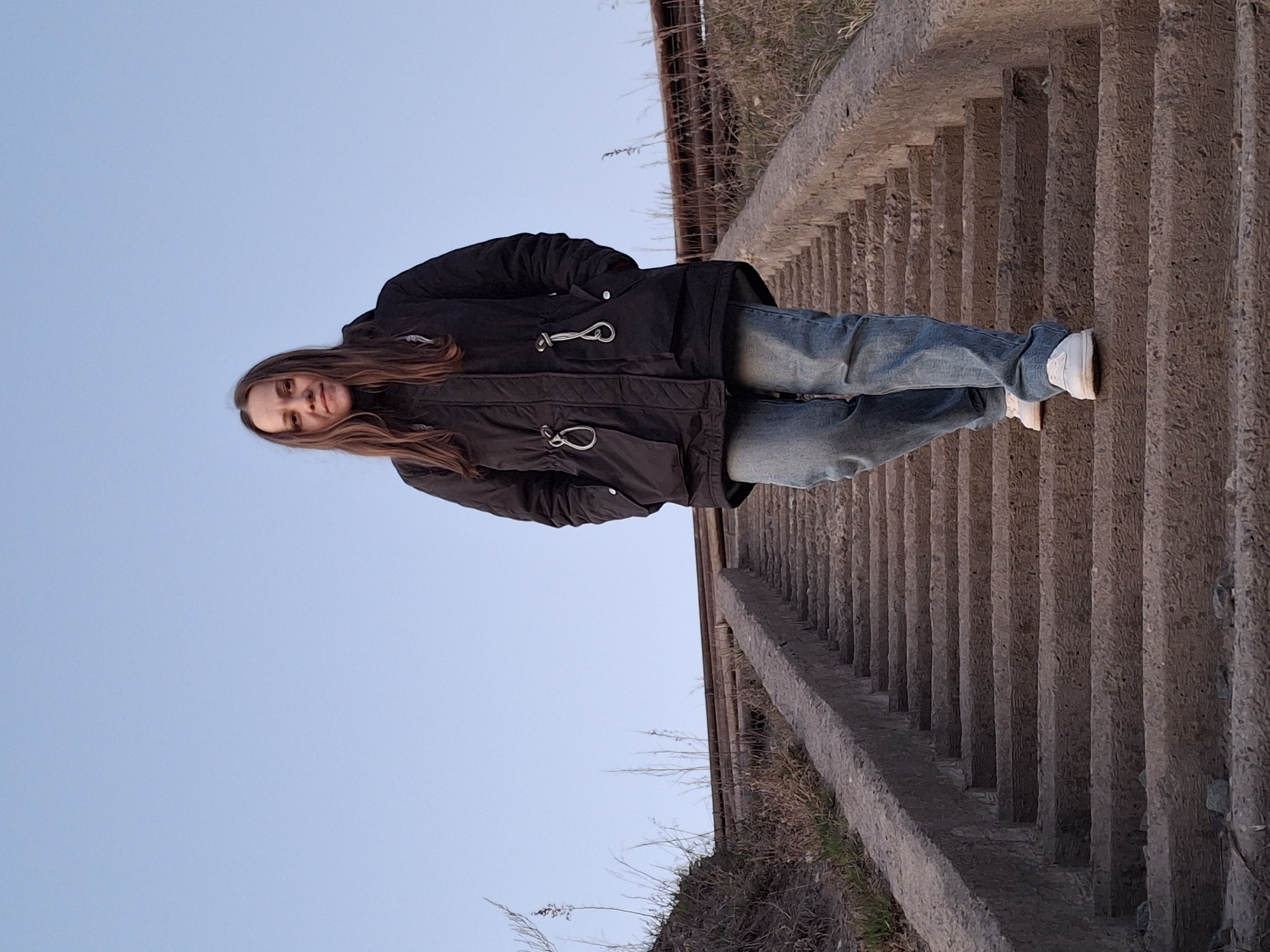 A woman stands on concrete steps under a blue sky. She wears a black jacket, blue jeans, and white sneakers. The steps are alongside a weathered concrete wall, with vegetation visible at the base. Above the steps is a metal railing and brown underbrush. The image captures a casual, outdoor scene, likely in Chaikovsky, Russia, though specific landmarks or identifiers are not directly visible in the frame.