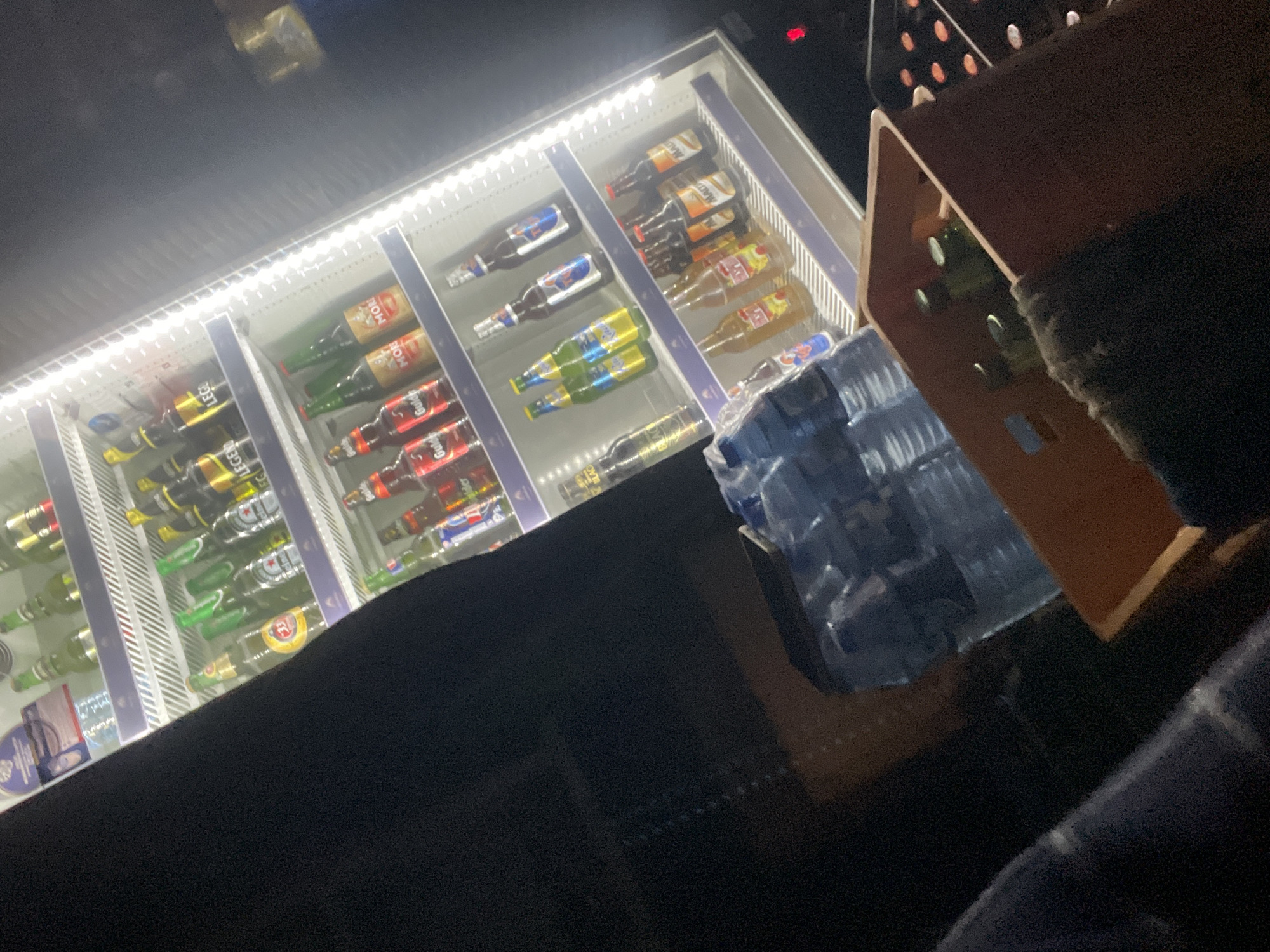 The image captures a dimly lit scene inside a refrigerated display case filled with various beverages, primarily bottles of beer and water. The display case, illuminated by internal lights, is tilted at an angle. Rows of bottles are neatly arranged on shelves. Prominent brands include "Legend," "Gulder," and what appears to be "Malt." The bottles vary in color, with many being green, brown, and clear glass. To the right of the display, a crate filled with green-bottled drinks is visible. In the foreground, a person's dark clothing is partially visible on the left side of the image, suggesting they are standing in front of the display. The overall atmosphere is dark, indicating it might be nighttime or an indoor setting with minimal ambient light. The presence of a refrigerated display case stocked with beverages suggests a commercial establishment, possibly a bar, restaurant, or retail store. The mention of Jos, Nigeria, in the prompt implies that this scene is situated in that specific location, although there are no explicit visual cues within the image itself to confirm this geographical detail.