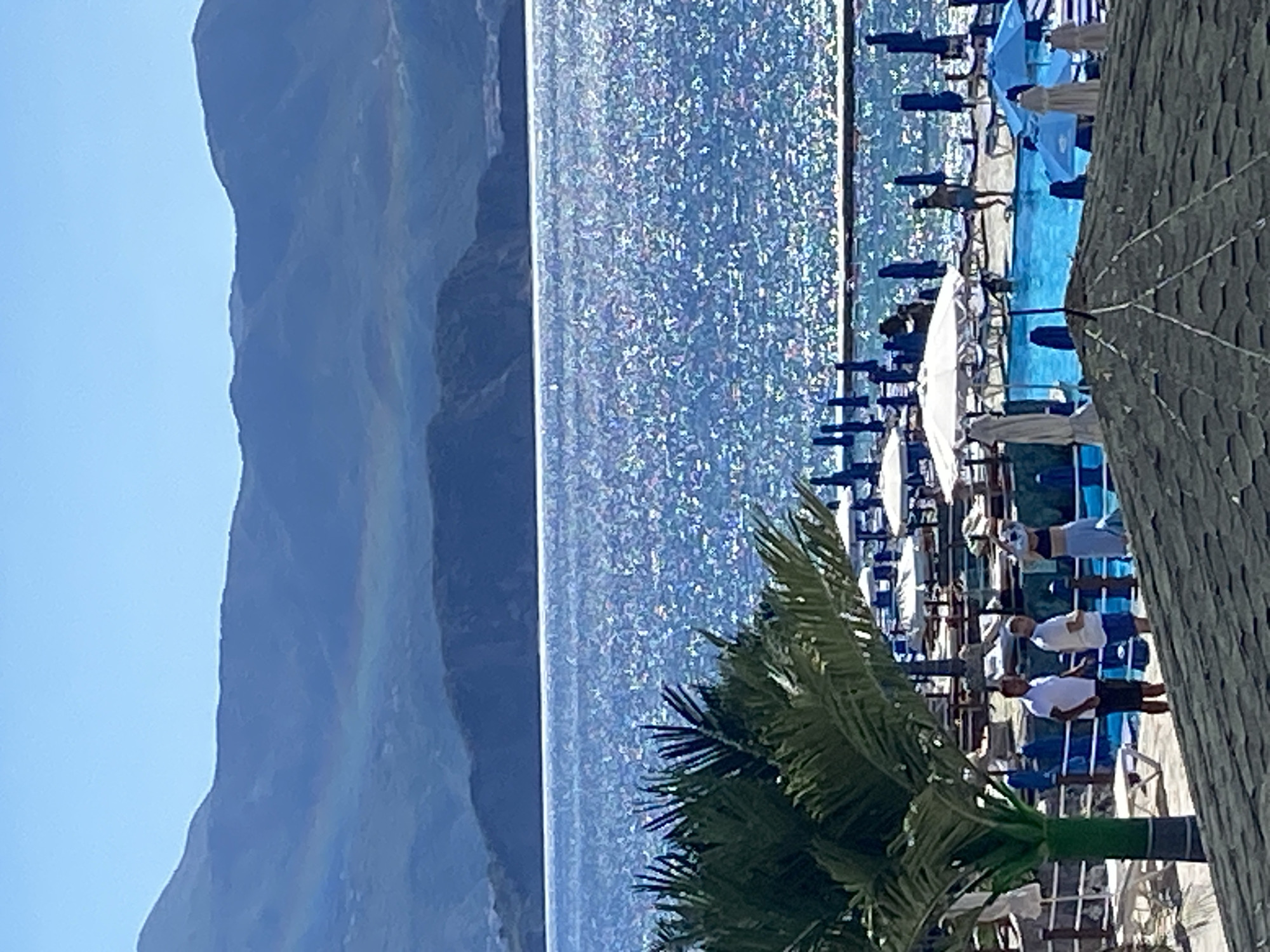 In Prijevor, Montenegro, a bright, sunny day unfolds at a coastal resort. The scene is dominated by the shimmering, sun-drenched Adriatic Sea, its surface a mosaic of sparkling light. In the foreground, a lush green palm tree stands tall, its fronds reaching towards the sky. Beyond it, a resort area unfolds with a vibrant blue swimming pool, surrounded by lounge chairs and umbrellas. Several people are visible enjoying the leisure facilities: some are standing near the pool, while others are interacting in or around the water. In the distance, majestic, layered mountains rise, their slopes shrouded in a hazy, atmospheric perspective, with a faint rainbow arching across them. The sky above is a clear, brilliant blue, completing the picturesque view of a tranquil vacation spot.