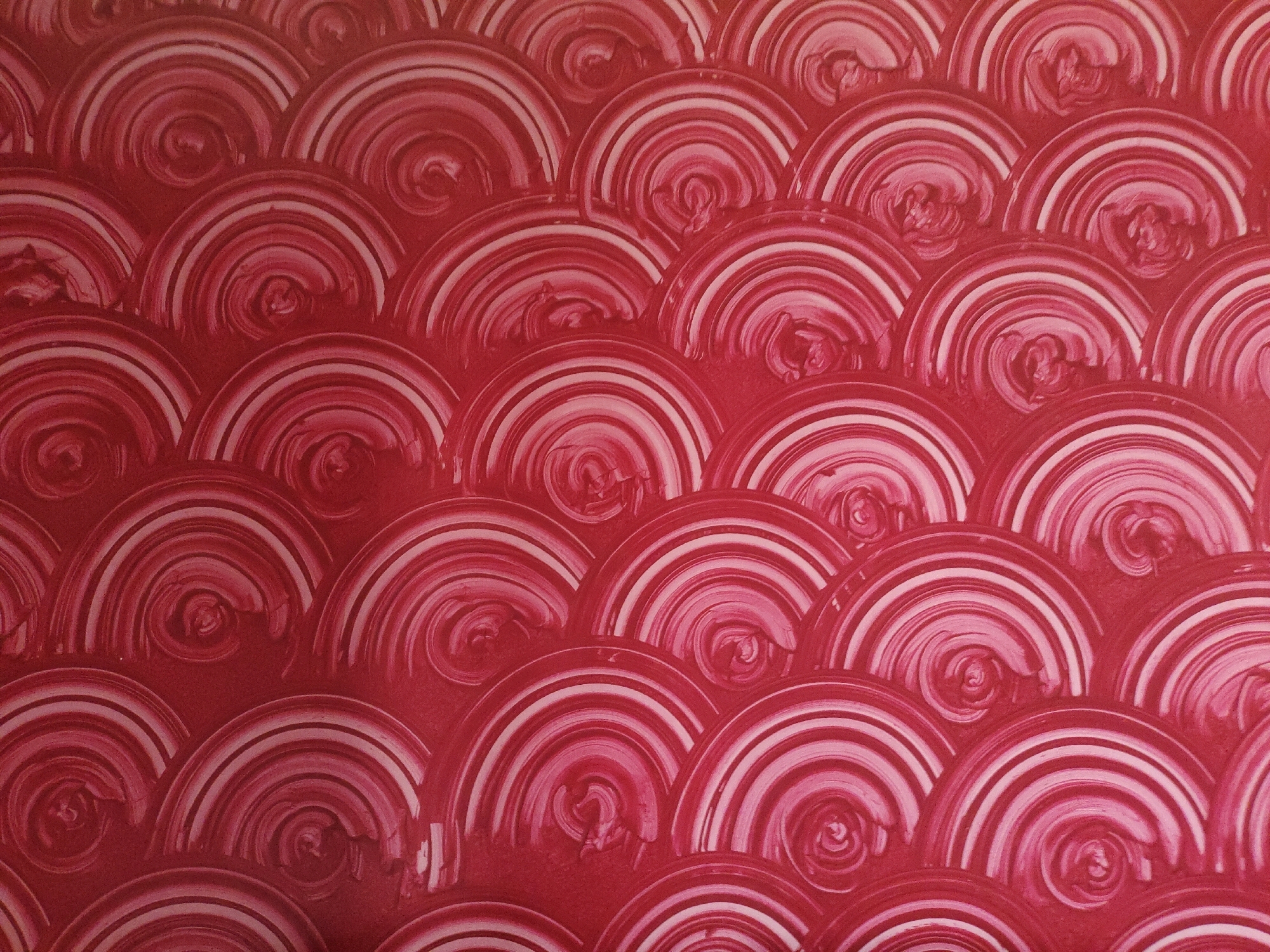 The image is a close-up, full-frame shot of a red textured wall with a repeating pattern. The pattern consists of semi-circular shapes, each with concentric rings of slightly varying shades of red and white, creating a swirling effect within each semi-circle. The texture of the wall appears to be a stucco or plaster finish. There are no people, objects, or any indication of a specific location within the city of Rampuram, India. The image solely focuses on the wall's surface and its decorative pattern.