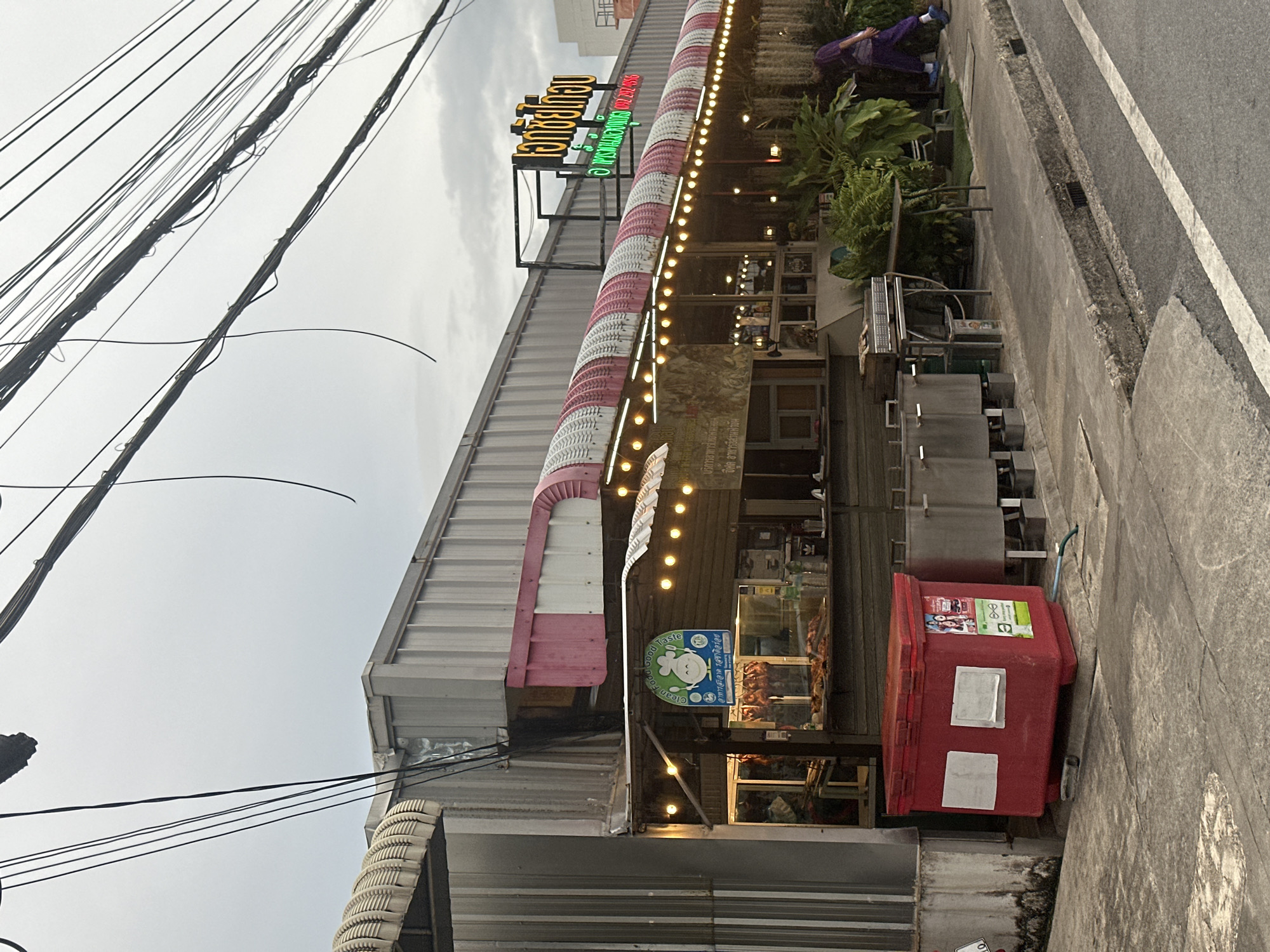 The image depicts a street-level view of a food stall or small restaurant in Phuket City Municipality, Thailand. The establishment has a corrugated metal exterior with a distinctive pink and white striped awning illuminated by a string of yellow lights. Several signs are visible, including one with Thai script and a cartoon chicken graphic that reads "Clean Food Good Taste." In front of the stall, there are metal food preparation stations and a large red cooler. A person wearing purple clothing is walking away from the stall towards the right side of the frame. The street in front of the establishment appears to be paved, with a concrete sidewalk and a drainage ditch. Overhead, a tangle of electrical wires crisscrosses the sky, which is overcast. The scene suggests a commercial area, possibly a local market or a street with small businesses.