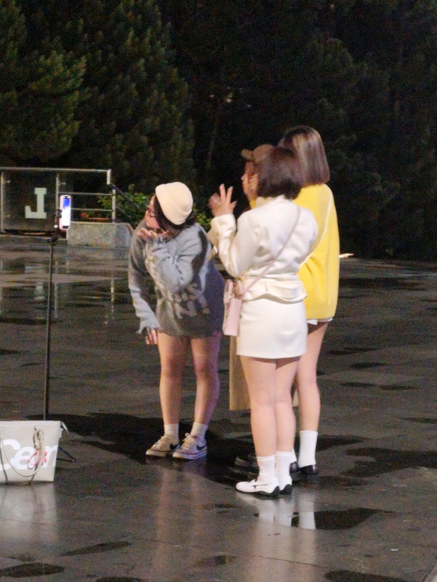 Four young individuals are gathered outdoors on a wet, reflective surface, possibly a street or plaza at night. Three are women, and one is a man. One woman, wearing a light-colored beanie and a grey sweatshirt dress, is leaning forward and blowing a kiss towards a smartphone mounted on a tripod. To her right stands another woman in a white blazer and skirt, with a pink crossbody bag. Behind her, a third woman wears a bright yellow sweater. The man, partially obscured, is behind the woman in white. The background features dark trees and what appears to be a large, illuminated "J" sign. The overall atmosphere suggests a casual social gathering or perhaps a live stream event. The scene is located in Da Lat, Vietnam.