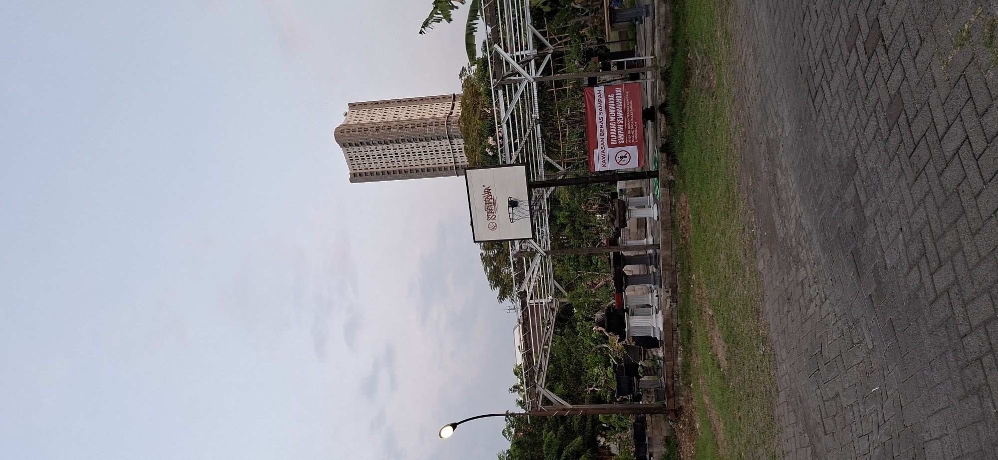A tall, slender, beige apartment building with many windows stands against a pale, overcast sky. In the foreground, a white basketball backboard with a red logo and net is visible. The backboard is mounted on a black pole. To the left of the basketball backboard, a street lamp with a glowing light is positioned. Behind the basketball backboard, a covered structure made of metal poles and beams supports a canopy or roof. This structure is over a display of potted plants, many of which appear to be bonsai trees, resting on decorative pedestals of various sizes and colors. A red sign with white text is attached to the pole of the basketball backboard, reading "KAWASAN BEBAS SAMPAH" and "DILARANG MEMBUANG SAMPAH SEMBARANGAN!" with a symbol of a person throwing trash into a bin with a line through it. The ground in front of the structure is paved with dark grey bricks arranged in a herringbone pattern, and beyond that is a strip of green grass. The overall scene suggests an outdoor area, possibly a park or public space, with a blend of recreational and decorative elements, and a residential building in the background.