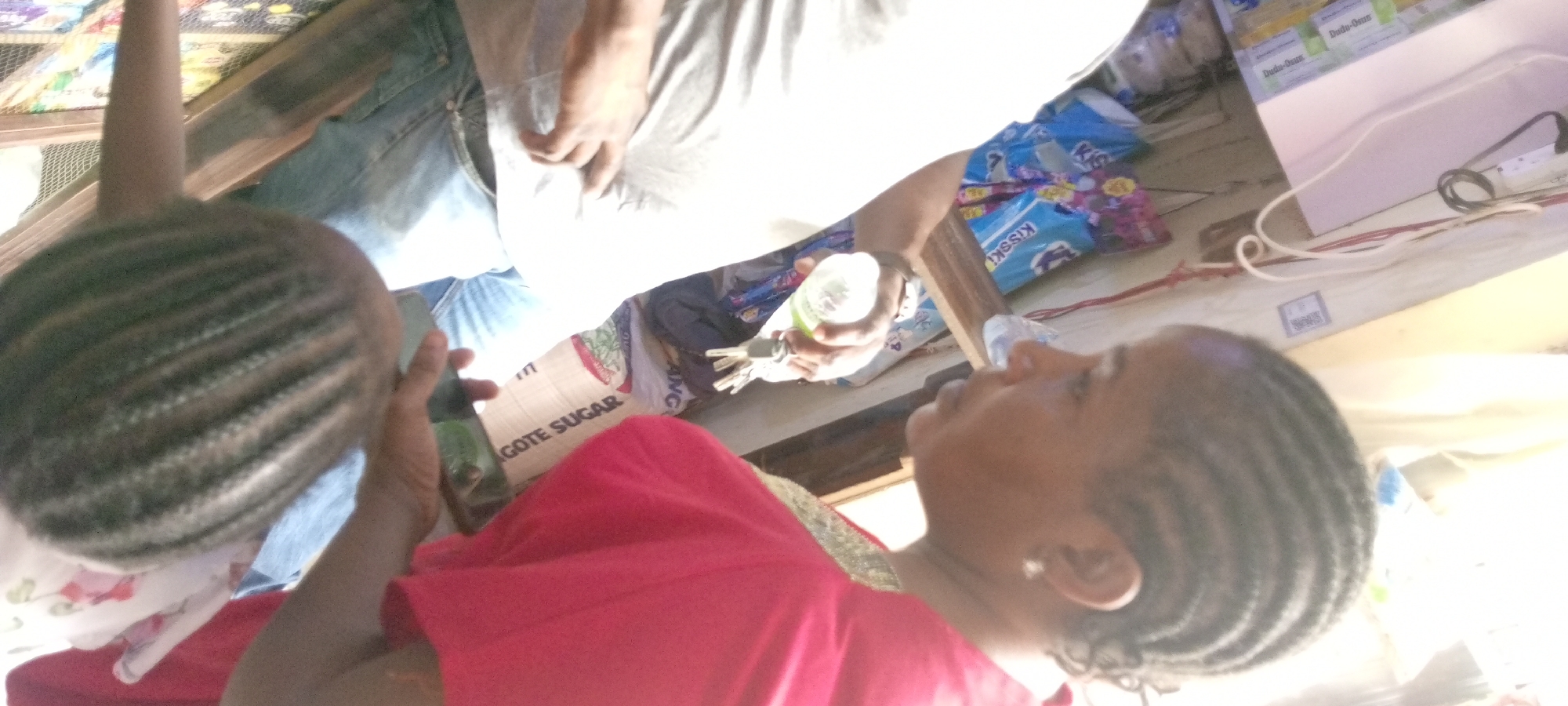 The image, taken in Gayawa, Nigeria, captures a sideways view of an indoor or semi-outdoor setting, likely a small shop or market stall. The scene features three individuals and various packaged goods.
In the foreground on the left, a child's head is visible, adorned with dark cornrow braids. The child wears a light-colored top with a floral pattern. To the right, an adult female is seen in profile, looking towards the left. She also has cornrow braids and is dressed in a vibrant red garment, possibly with green trim. A small earring is visible on her right ear.
Towards the center, partially obscured by the woman, another individual (likely male) is visible from the waist down, wearing blue jeans and a light-colored t-shirt. This person holds a small, clear plastic bottle with a white cap in one hand and a bunch of keys in the other.
The setting includes several commercial products. A large white bag prominently features the text "DANGOTE SUGAR". Nearby, blue packages with "KISSKISS" branding are visible. On a white surface or shelf to the right, small boxes are stacked, bearing the label "Dudu-Osun". White and red electrical cables are also visible on a surface in the background, alongside some wooden structures that may be shelving.
The activity appears to be an everyday interaction or transaction taking place within the shop. The bright lighting suggests it is daytime. The presence of specific branded products like Dangote Sugar, Kisskiss, and Dudu-Osun serves as a strong location cue, consistent with the Nigerian context. Emotions are not clearly discernible from the partial views of the individuals.
