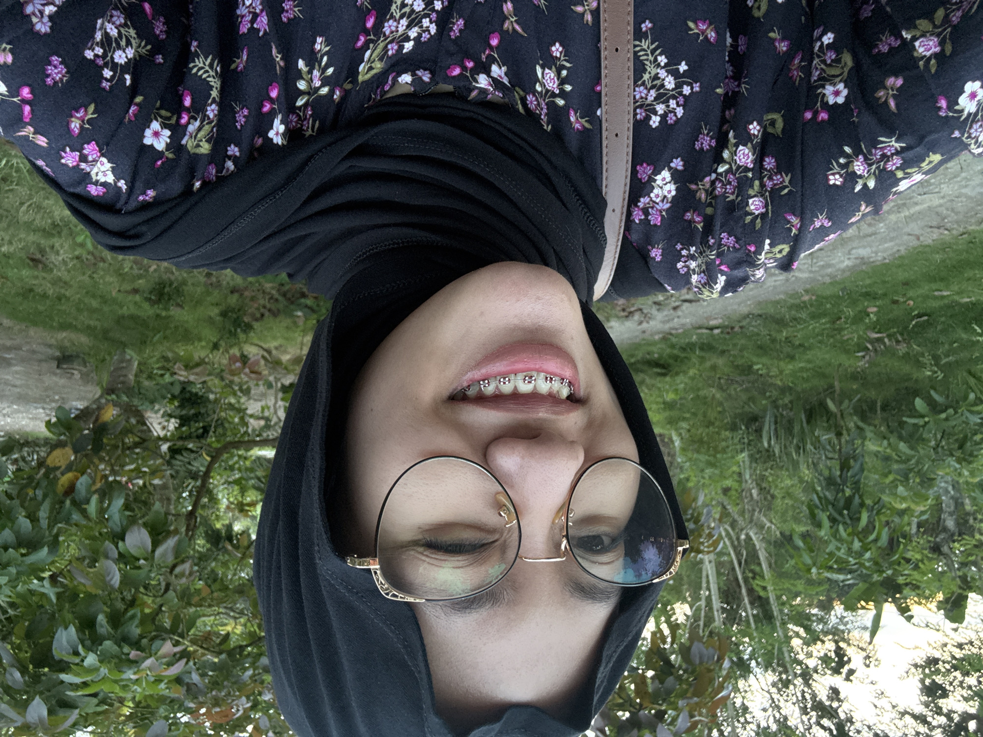 A woman with braces and round glasses is wearing a black head covering and a floral print shirt. The picture is taken from a low angle, with green foliage visible in the background. The location is likely Gadingrejo, Indonesia.