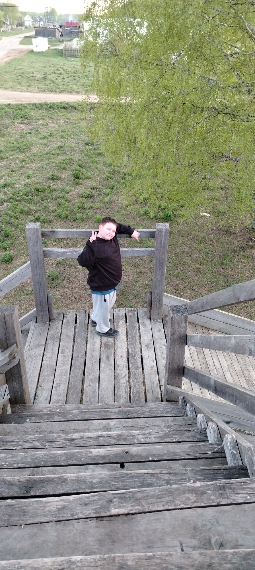 A medium shot taken from above shows a young person standing on a wooden tower. The tower appears to be built over a grassy field near the Russian city of Зипуново.
The person, who has lighter skin, wears a dark sweatshirt, grey pants, and black shoes. They are leaning against the wooden railing and holding up two fingers in a peace sign. The railing surrounds the top level of the wooden tower, which descends into a set of stairs. A tree with green leaves is situated to the right. The background features a rural scene with small buildings and a dirt road.