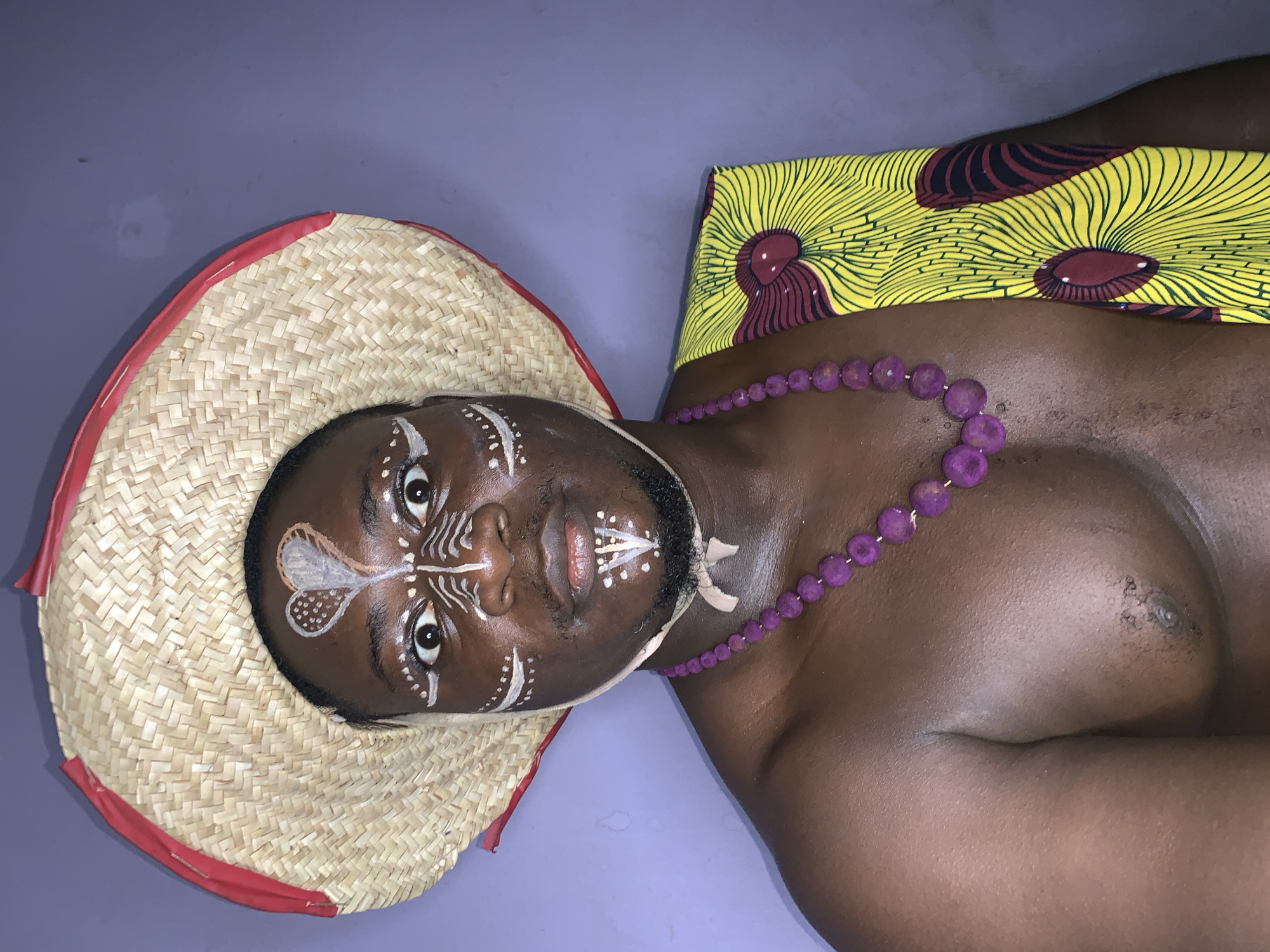 A portrait of a Black man is shown lying down on a purple surface. The man is wearing a woven straw hat with a red trim and has white face paint on his forehead, around his eyes and mouth, and on his chin. He is also wearing a necklace of large purple beads. To his right, the man is draped with a piece of fabric that has a yellow background with a repetitive pattern in burgundy and black. The fabric appears to be a vibrant African print. The man's skin is dark, and he has a beard and mustache. His eyes are open and looking directly at the camera. The overall lighting is soft, and the scene appears to be indoors. The location context is Akwanga, Nigeria.