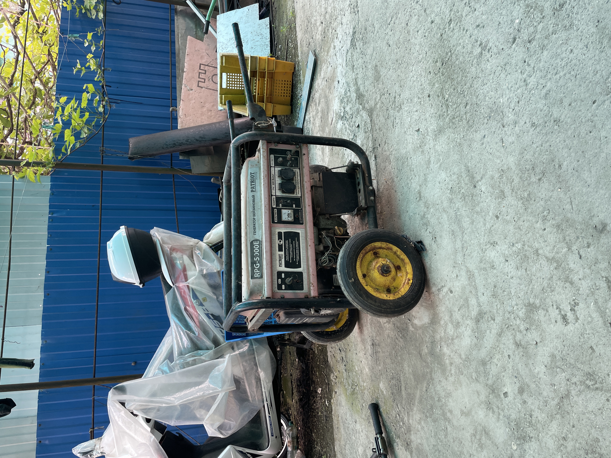 The image depicts a Patriot RPG-5000E gasoline generator on a concrete surface, possibly an outdoor area. It is surrounded by various items, including plastic sheeting, yellow crates, and a blue corrugated metal wall in the background. The generator appears to be used or stored, judging by its condition and the cluttered environment. Based on the OCR text, the generator is labeled in Russian, indicating it is likely located in Russia. The image does not provide any explicit evidence or information about it being in Olginka, Russia.