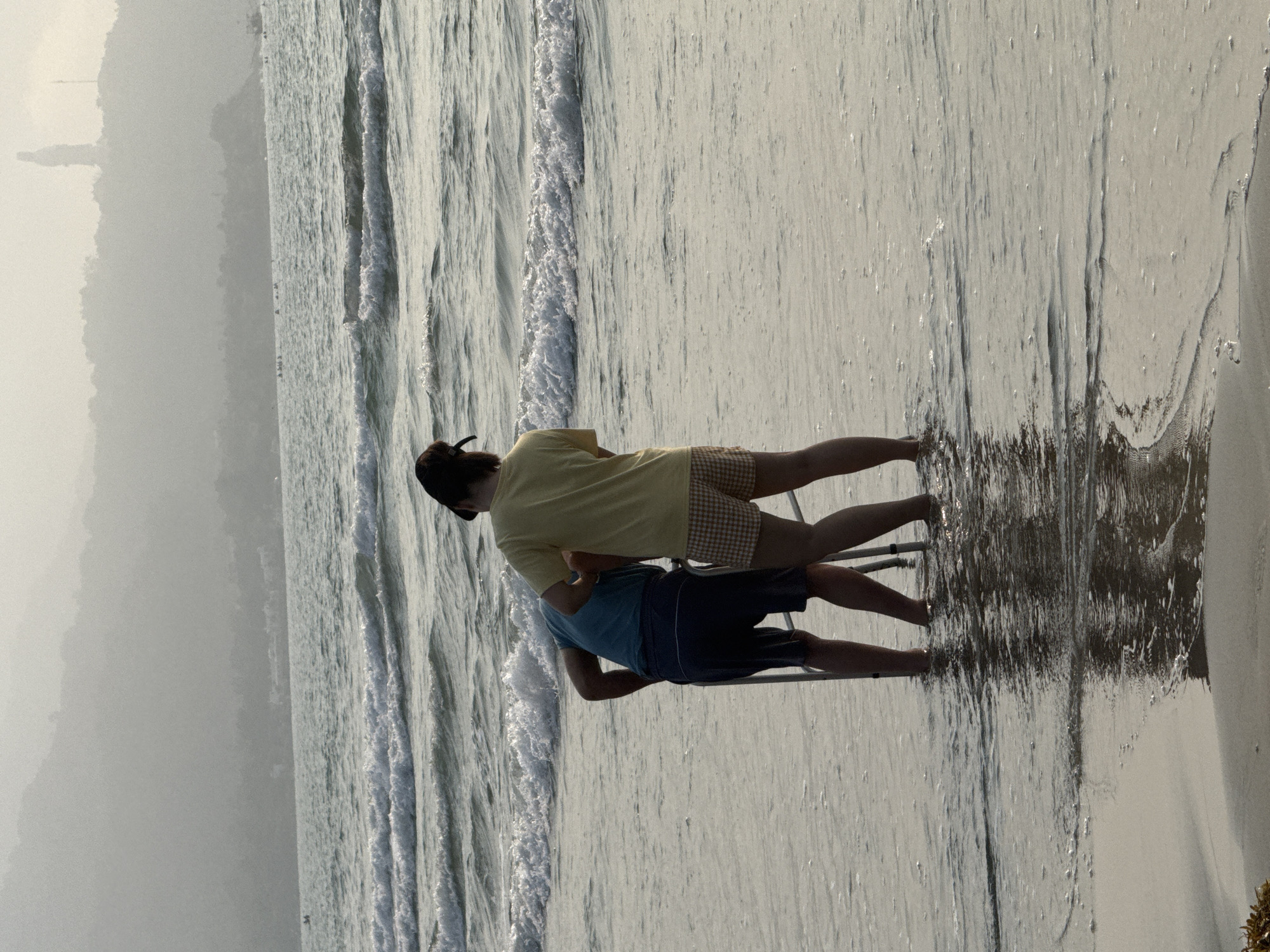Two people stand in the surf on a beach in Đà Nẵng, Vietnam. The person in the foreground wears a blue shirt and navy shorts, while the other wears a yellow shirt and brown plaid shorts. They are standing on or near a silver metal object (possibly a ladder or scaffolding) in the water. Waves roll onto the shore. Hazy mountains are visible in the distance. The sky is overcast.