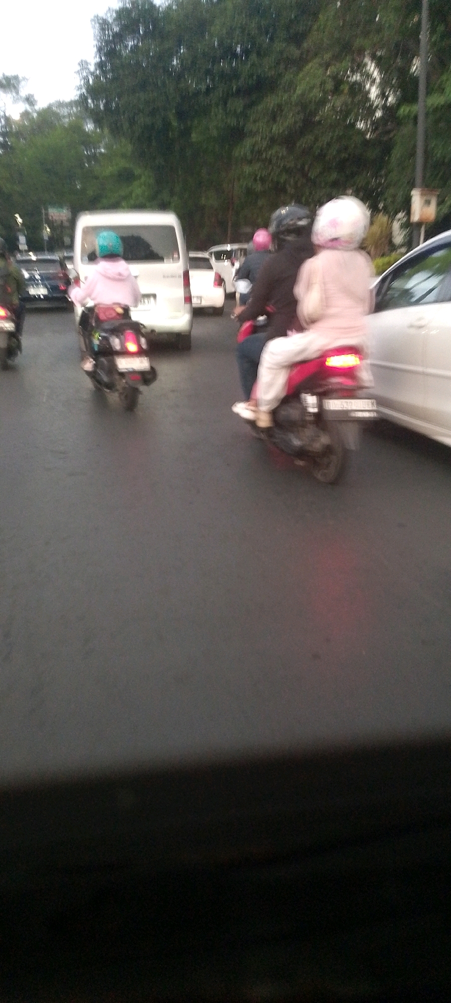 The image shows a traffic scene in what is identified as Bandung City, Indonesia. Two motorcycles are prominently featured in the foreground. The motorcycle on the left has a rider wearing a pink hoodie and a light blue helmet, while the motorcycle on the right carries two people, a driver wearing a black helmet and a passenger in a light pink outfit and a white helmet. Both motorcycles are in motion, as indicated by the motion blur. In the background, there are several cars and a white van. Trees line the street, suggesting an urban environment with greenery. The road appears wet, indicating recent rain or a humid climate. The lighting suggests it is either early morning or late afternoon. The overall scene depicts a typical urban traffic scenario.