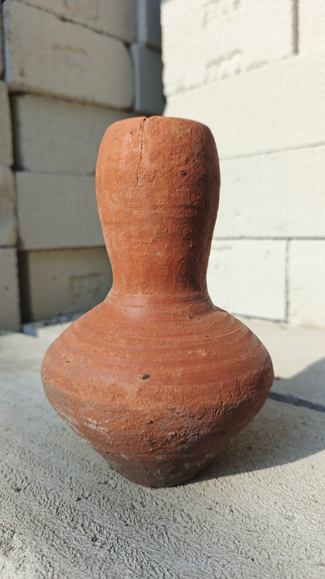 A terracotta pot with a narrow neck and a wide, rounded base is positioned in the foreground, casting a shadow on the textured concrete surface it rests upon. The pot appears weathered, with visible cracks and imperfections on its surface, and is a reddish-brown color. In the background, a stack of pale, rectangular bricks is visible, some with faint markings. The scene is illuminated by natural light, suggesting an outdoor setting, possibly in a construction area or a place where materials are stored. The overall impression is one of simple, utilitarian objects in an outdoor environment.