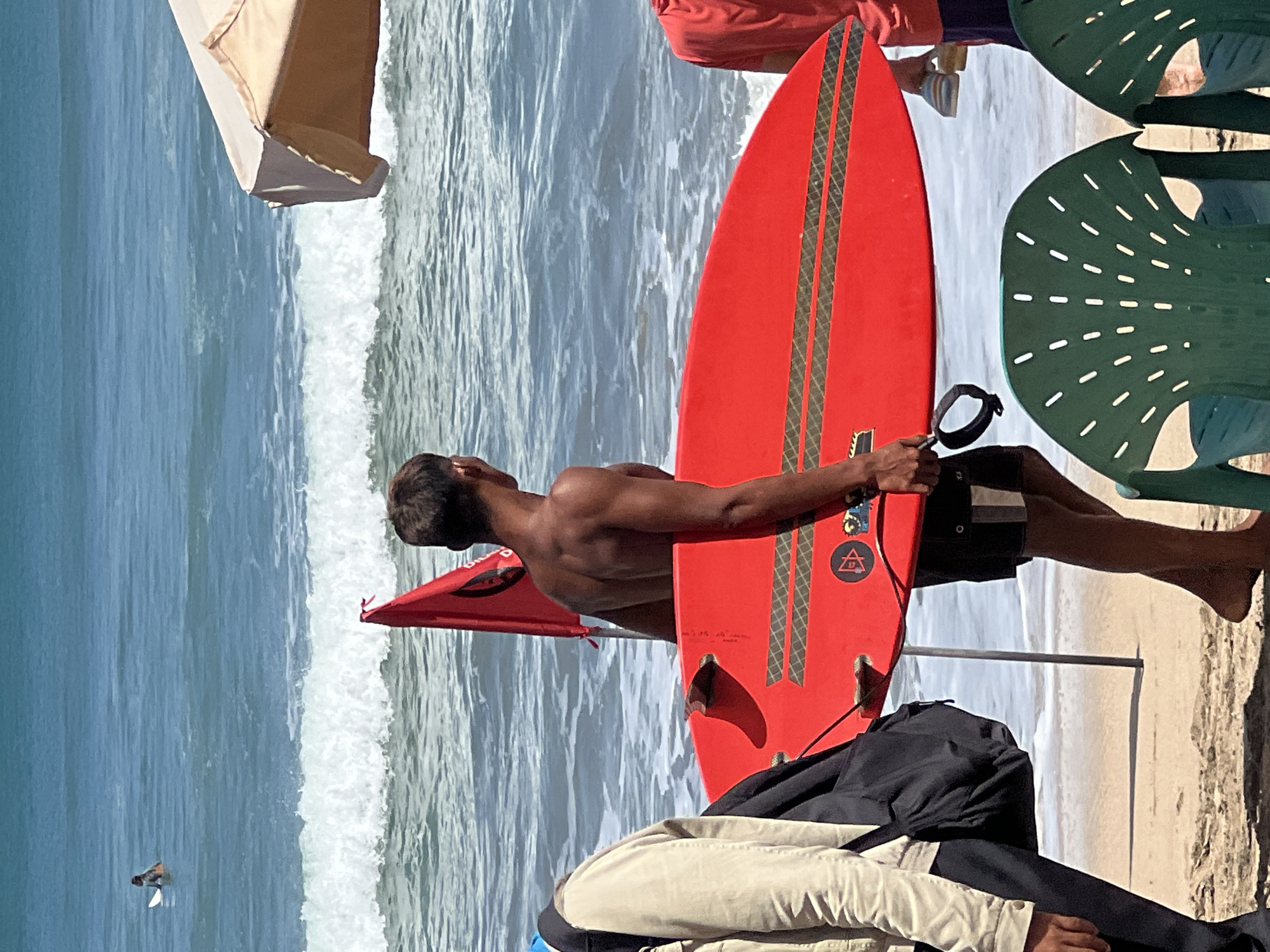 A surfer, carrying a bright red surfboard, walks on a sandy beach in Kuta, Indonesia, with the ocean waves in the background. The surfer is wearing dark shorts and appears to be walking away from the camera. A red flag is planted in the sand nearby. In the foreground, two green plastic chairs are visible, with portions of other people and a beach umbrella also in view.