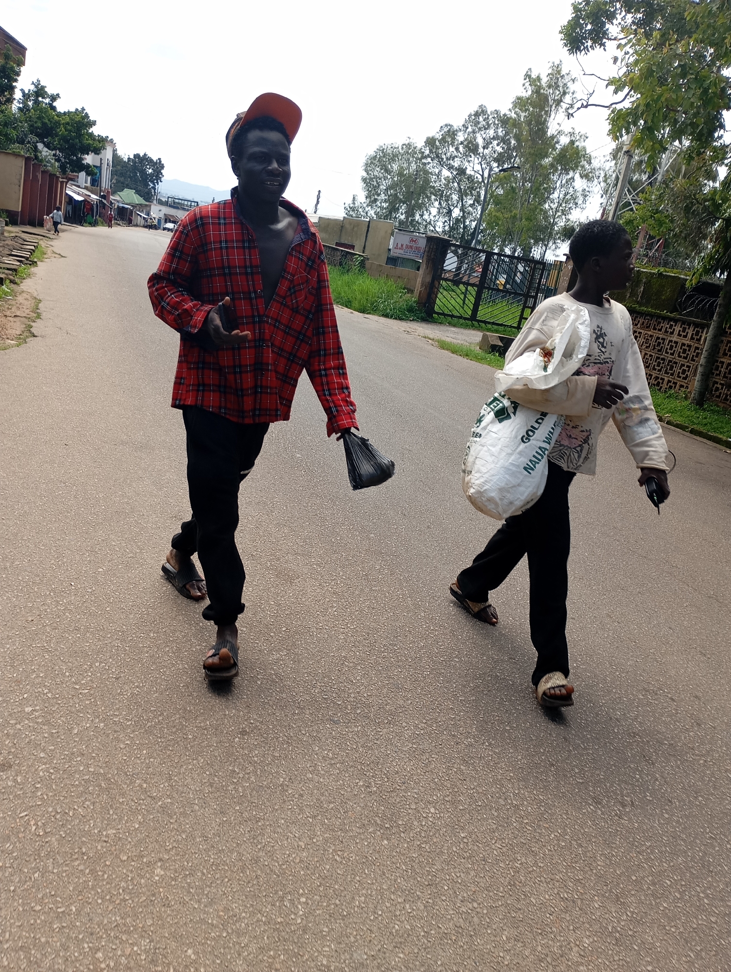 Two individuals are walking on an asphalt road in what appears to be a moderately urban area of Jos, Nigeria. The individual on the left, closer to the viewer, is a dark-skinned male wearing a red and black plaid open shirt, dark pants, and sandals. He has an orange cap on backwards and is holding a black plastic bag in his right hand. The individual on the right is a lighter-skinned individual wearing a light-colored long-sleeved shirt with a graphic print and dark pants. They are carrying a large white woven bag over their left shoulder and holding a dark object, possibly a phone, in their right hand, also wearing sandals. The road is bordered by a sidewalk with some vegetation and buildings on the left, and a fence and more trees on the right. The sky is overcast. The context suggests a daily scene in Jos, with people going about their business.