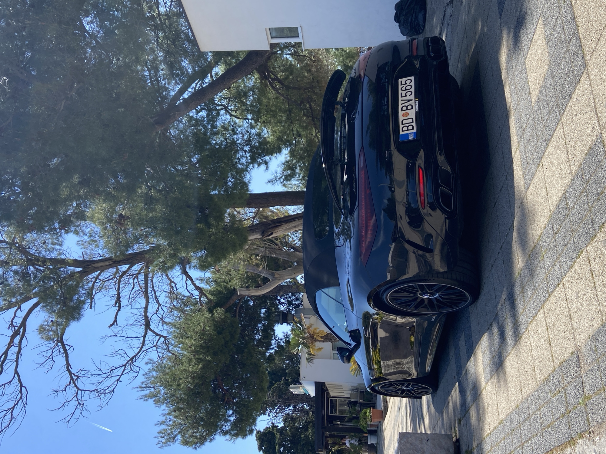 A black convertible car is parked on a paved surface. The car's license plate reads "BD BV565" and indicates it is from Montenegro ("MNE"). The car is parked near a building with white walls and windows. Several trees with green foliage are also present. The sky is visible and blue. The location appears to be in Bar, Montenegro.