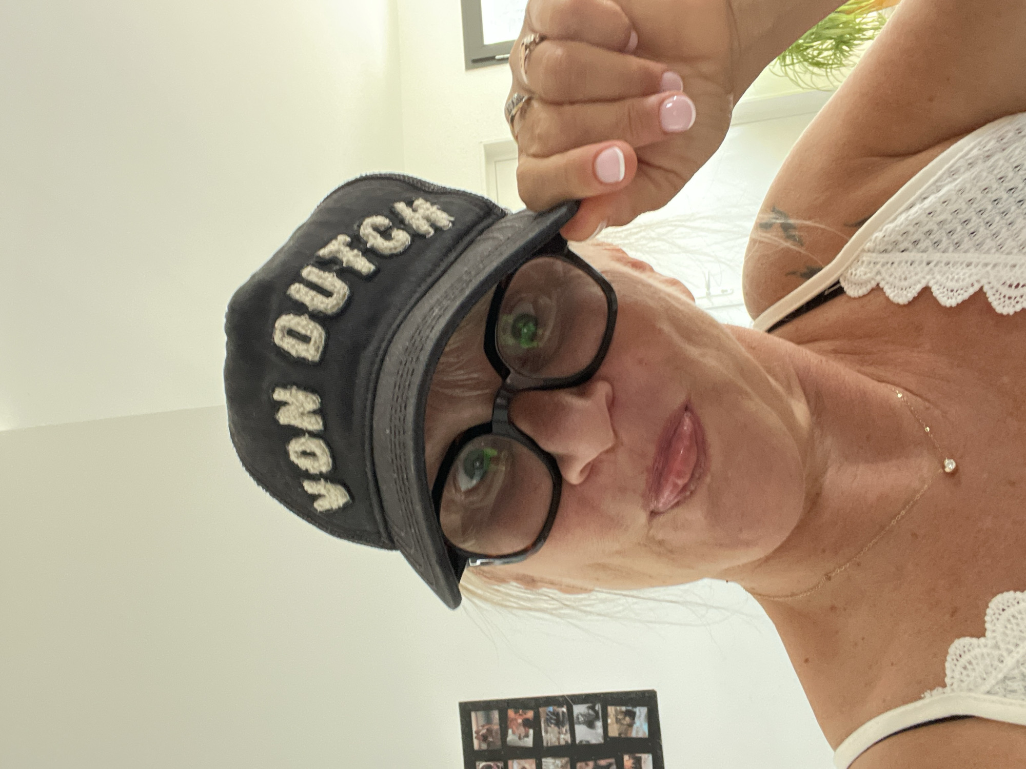 In an indoor setting, a woman with blonde hair, wearing black-rimmed glasses and a black baseball cap with "VON DUTCH" embroidered on it, sticks her tongue out at the camera. She is also wearing a white lace-trimmed top and a delicate gold necklace with a small diamond pendant. Her right hand, adorned with rings, is lifting the brim of her cap. To her left, a framed collage of small photographs is visible on a light-colored wall. The background suggests a home environment with soft lighting. There is no identifiable location in Chambœuf, France.