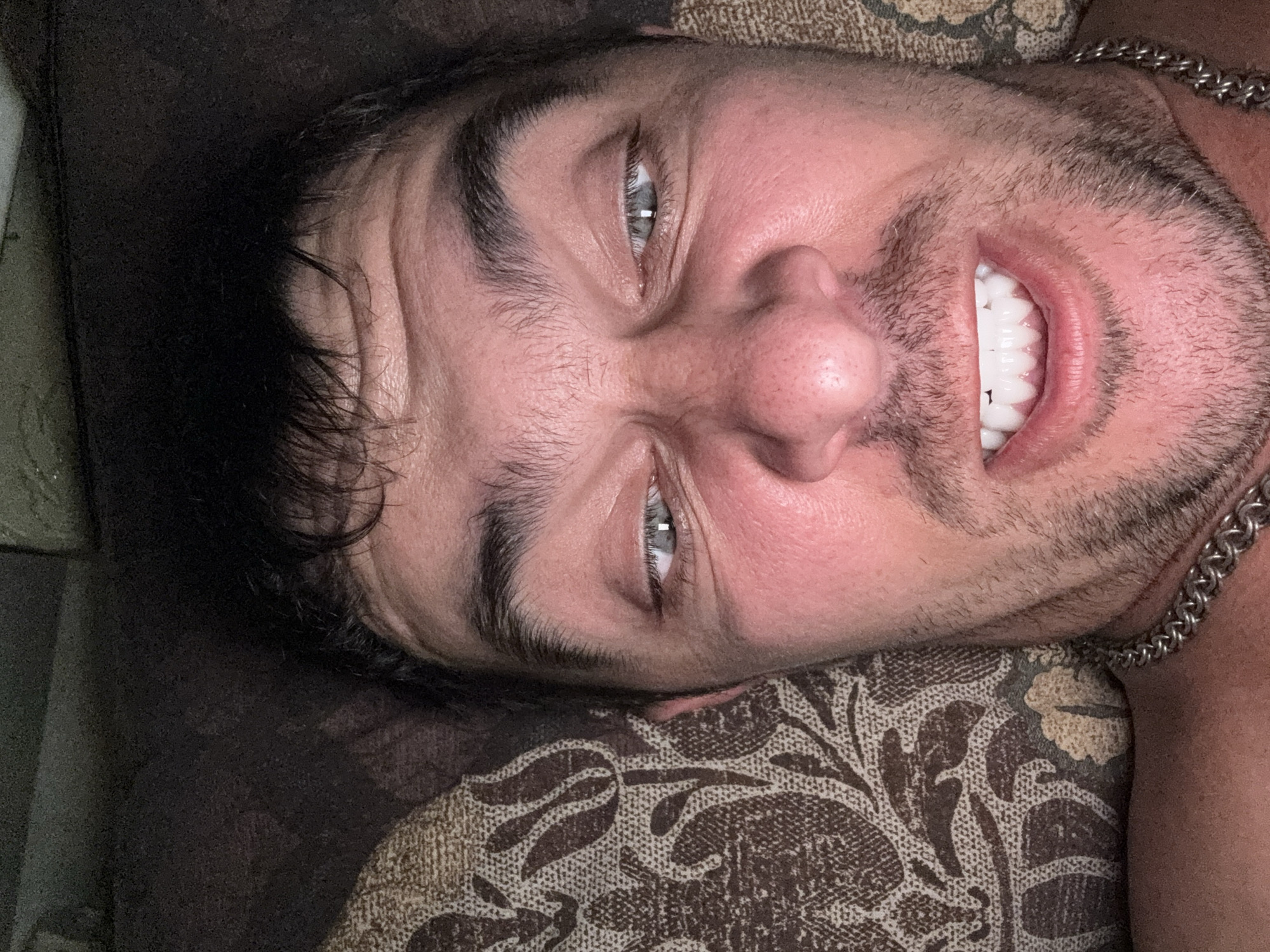 The image is a close-up, overhead shot of a man's face, taken indoors. The man has dark, messy hair, thick eyebrows, and light blue eyes. His face is flushed, and he has a noticeable amount of stubble on his chin and jawline. His mouth is open, revealing white, even teeth, and he is wearing a thick silver chain around his neck. He is lying down, as indicated by the patterned fabric of a cushion or blanket behind him and to his left. The lighting is dim, suggesting it might be nighttime or a dark room. No specific location in Shakhty, Russia is identifiable from the image.