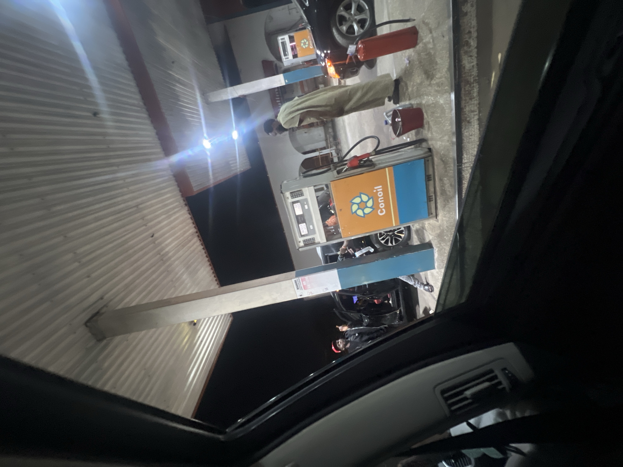 The image depicts a Conoil petrol station at night. A person in a long, light-colored garment is standing near a fuel pump, possibly attending to a vehicle. Another person, wearing a dark jacket and a red hat, is near a different car and fuel pump. Buckets are visible near the pumps. The photograph is taken from inside a car, looking out through the sunroof. It is not possible to verify the specific location within Abuja without more information.