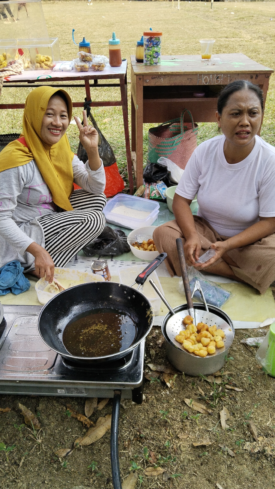 Two women are preparing food outdoors in what appears to be Margatunggal, Indonesia. The woman on the left, wearing a yellow hijab and striped pants, smiles at the camera while holding up two fingers in a peace sign. She is dipping dough into a batter in a white plastic container. In front of her is a small portable gas stove with a frying pan containing hot oil. The woman on the right, wearing a white t-shirt and brown pants, looks towards the camera with an expression of surprise or concern. She is holding a small plastic container. In front of her, a metal strainer filled with fried dough pieces sits in a larger metal pot. Other food items and containers are scattered around them, including bottles of sauces, a plastic cup with a drink, and a large plastic bin filled with what looks like flour. The scene is set on a patch of dry grass with scattered leaves and a few small green plants. In the background, there are two tables and some baskets. The overall impression is of an informal outdoor food preparation activity.