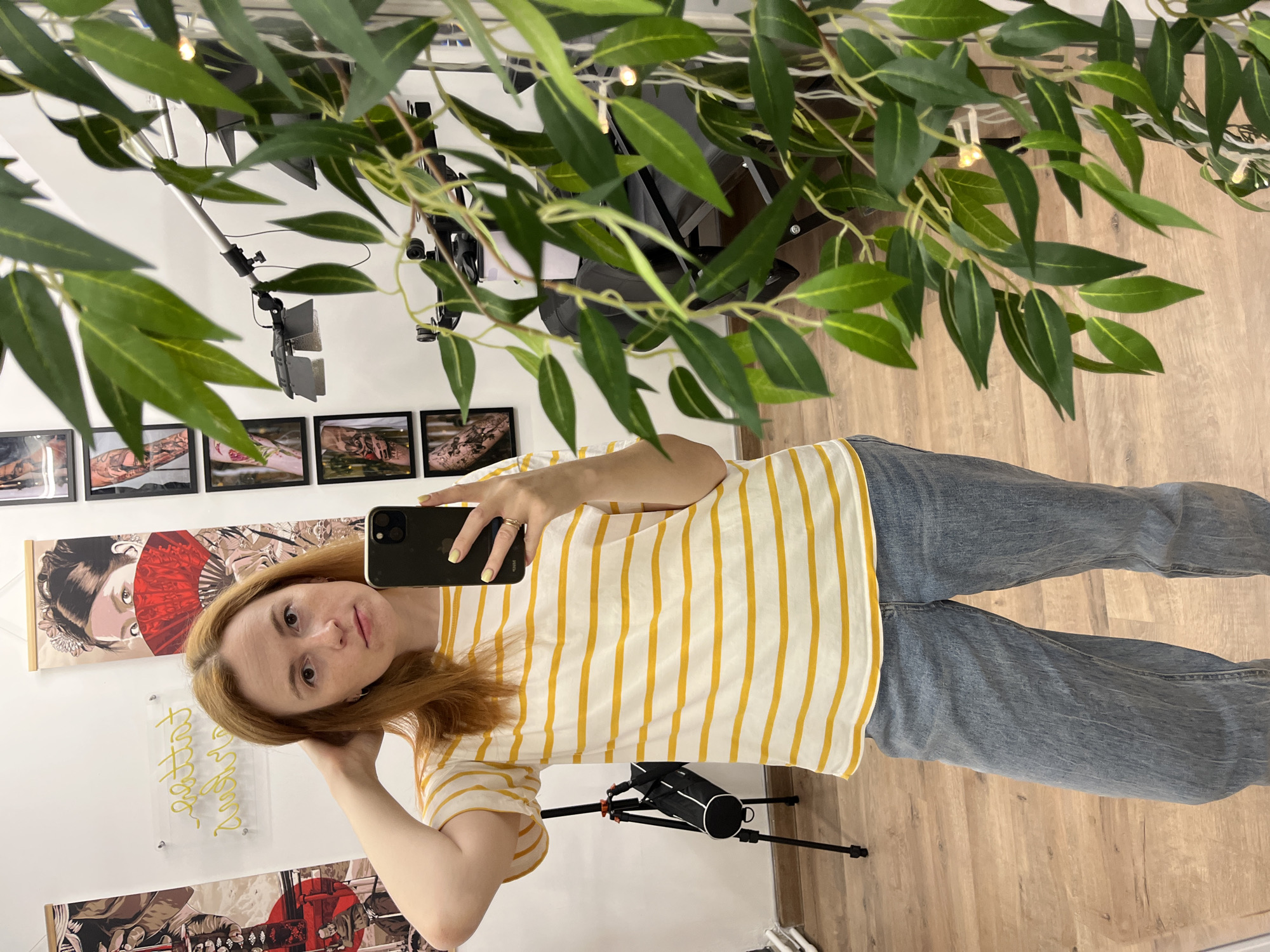 A person is standing in what appears to be a room, possibly a living space or a studio. The person is holding a black smartphone, presumably taking a selfie or a photograph. They are wearing a white t-shirt with yellow vertical stripes and blue jeans. The person has reddish-brown hair and is looking directly at the camera. Behind the person, a white wall features framed artwork and possibly an illuminated transparent sign. A plant with long green leaves is partially obstructing the view. There's also photographic lighting equipment and a tripod. The floor is wooden. The location appears to be indoors, possibly in the city of Chaikovsky, Russia, given the user's request.