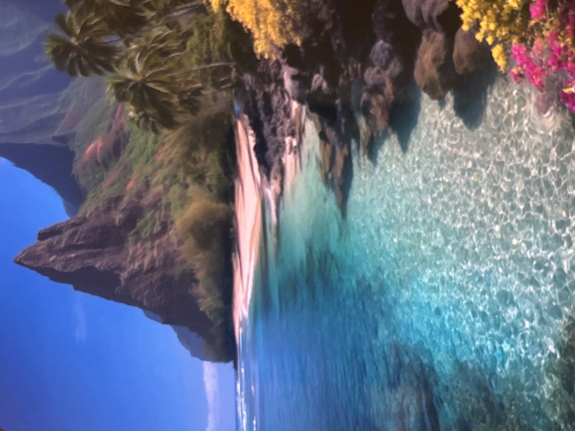 This image captures a breathtaking tropical coastal landscape, seemingly rotated 90 degrees counter-clockwise from its intended orientation. If viewed correctly, a clear, deep blue sky occupies the upper left quadrant. A majestic, steep, dark rocky mountain range or cliff dominates the left side and background, its slopes covered in rich, diverse green vegetation, with some areas showing reddish-brown earth.
Along the coastline at the base of these mountains, a narrow strip of light-colored sandy beach meets incredibly clear, sparkling turquoise to deep blue water. The water exhibits vibrant patterns of sunlight reflecting off its surface, indicating shallow depths near the shore and exceptional clarity. On the right side of the image, where the mountains meet the coast, lush yellow foliage and bright pink or magenta tropical flowers bloom vibrantly, adding a striking burst of color to the scene. Palm trees with their distinctive fronds are visible near the top of the mountain range on the left.
The scene depicts a serene, uninhabited natural environment. There are no people, buildings, or discernible objects, suggesting a pristine and untouched location. The bright illumination, clear sky, and sparkling water indicate it is daytime, likely under a sunny sky. The combination of dramatic cliffs, sandy beach, vibrant foliage, and crystal-clear water strongly suggests a tropical island or coastal paradise, possibly in the Pacific. No visible text is present in the image.