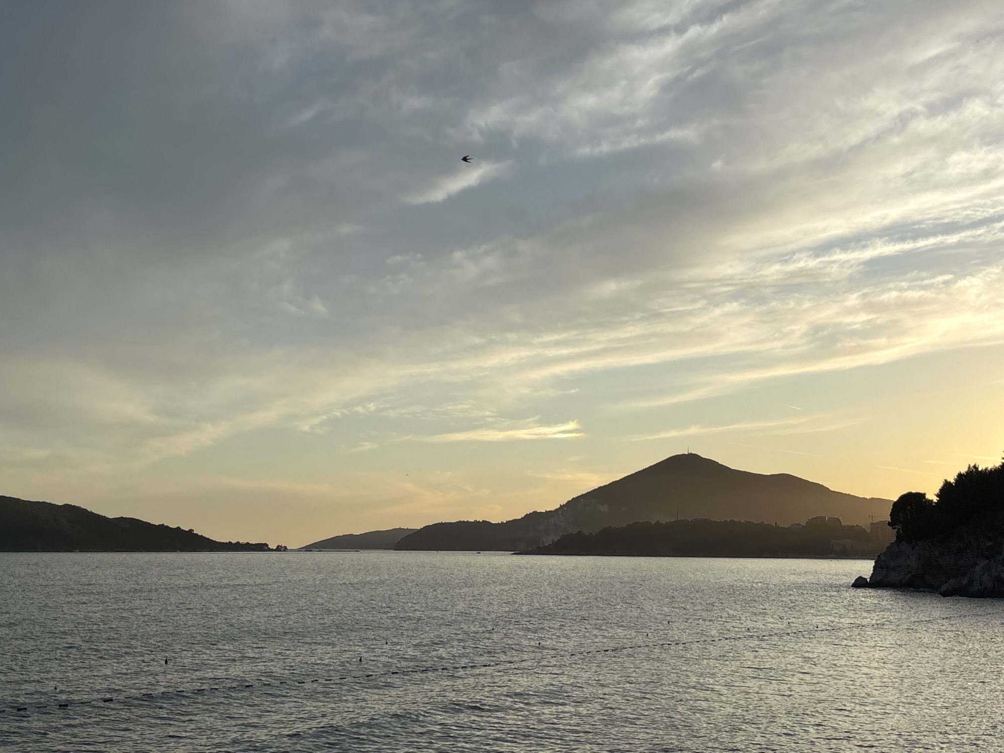 A serene seascape at dusk in Kamenovo, Montenegro, features the calm, dark water of the Adriatic Sea in the foreground. The sea is dotted with small buoys, forming a line parallel to the shore. In the middle ground, a series of silhouetted hills and mountains form a majestic backdrop, their peaks softly illuminated by the fading sunlight. A prominent mountain with a pointed peak stands out against the horizon. The sky above is a canvas of soft colors, with streaks of white and grey clouds interspersed with warm hues of yellow and orange, suggesting the last moments of sunset. A solitary bird is captured in flight against the vast sky. The shoreline is lined with dark vegetation, and on the right side of the image, a rocky outcrop juts into the sea. The overall atmosphere is tranquil and picturesque.