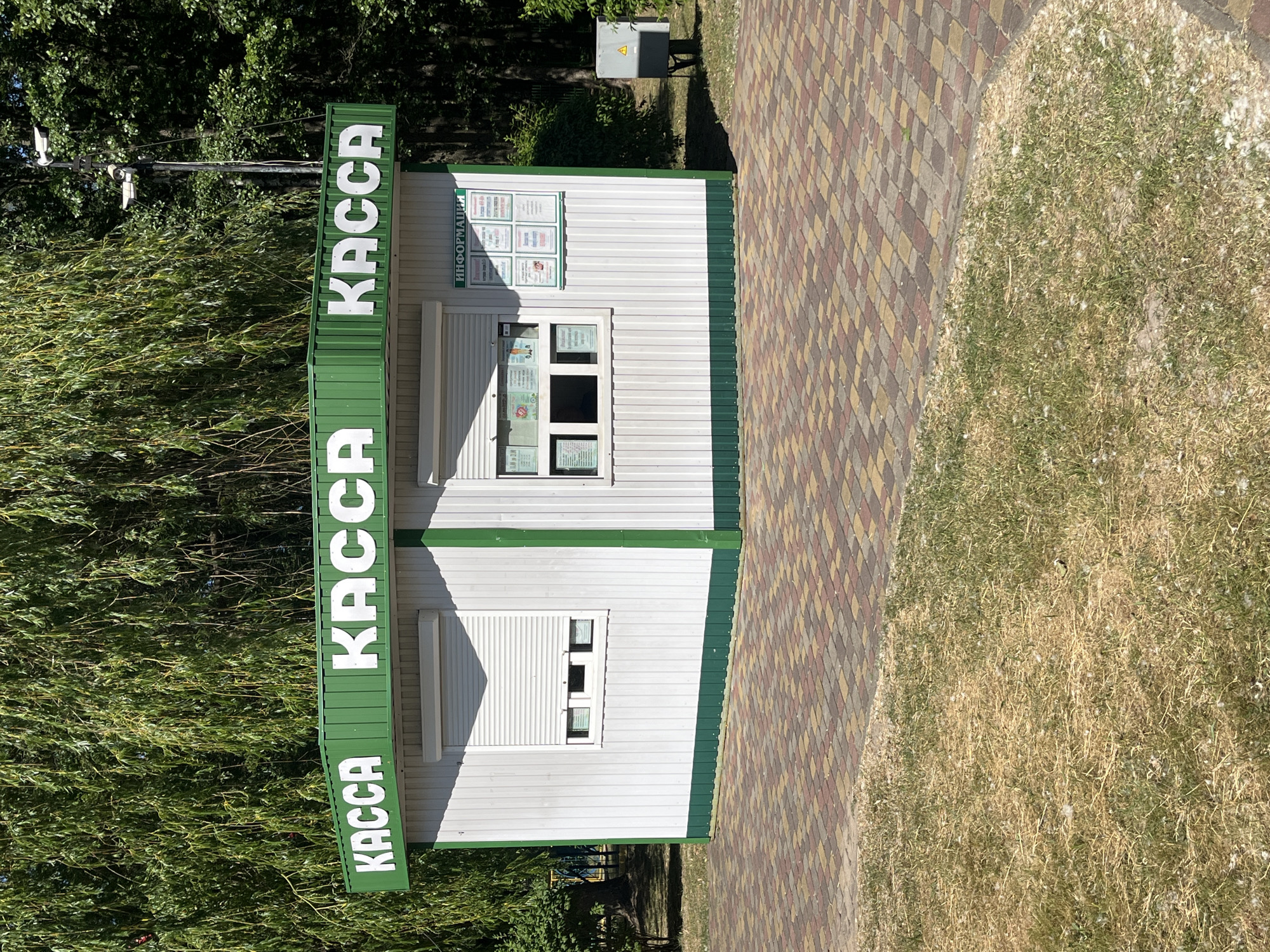 A white and green kiosk with the word "КАССА" (KASSA, meaning "cashier" or "ticket office") in large white letters on the green top edge is positioned in front of a lush green willow tree. The kiosk has two windows, one with a rolled-down security shutter and another partially open revealing what appears to be a menu or information display. The kiosk is situated on a paved walkway made of multi-colored square tiles. The area around the walkway consists of dry, yellowish grass. In the background, beyond the kiosk and tree, are more trees and a utility box with a yellow warning triangle. Two security cameras are mounted on a pole to the right of the kiosk. The scene is outdoors in what appears to be a park or recreational area.