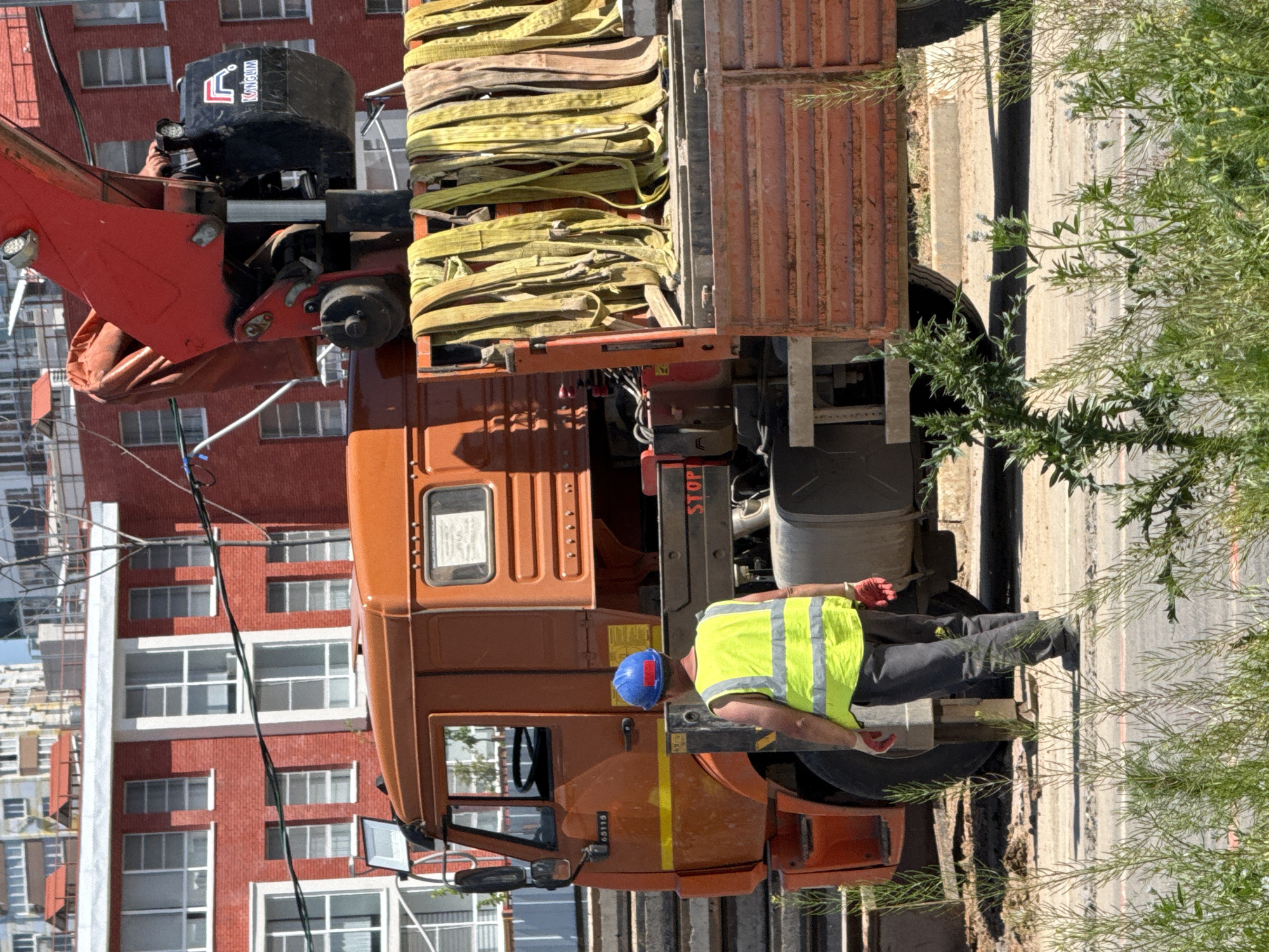 The image depicts a truck with a crane attachment on a street in what appears to be an urban area. A worker wearing a blue helmet and a bright yellow safety vest is near the rear wheels of the truck. The truck bed is loaded with multiple yellow straps. The truck is orange and has a red "STOP" sign visible. A multi-story brick building with numerous windows is visible in the background. Green foliage is present along the right side of the image. While I cannot definitively confirm the location as Rostov-on-Don, Russia, based on visual information alone, the presence of what appears to be Cyrillic text on the equipment suggests a location in Eastern Europe.