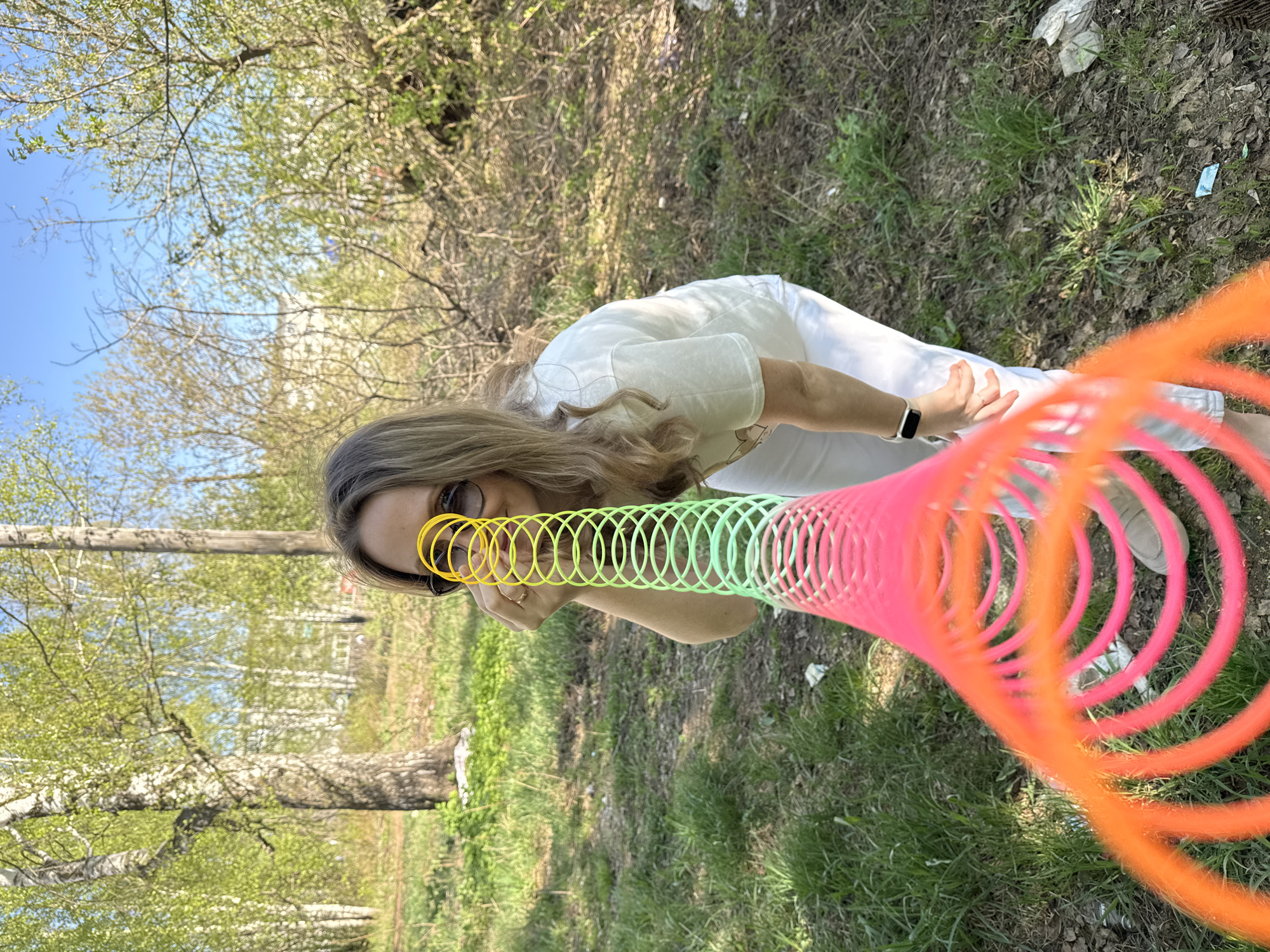 A young woman stands in a grassy area beneath trees, holding a colorful slinky toy that stretches across the frame from her face to the lower right. She wears sunglasses and light-colored clothing, including pants and a T-shirt. A watch is visible on her left wrist. The slinky is multicolored, featuring yellow, green, pink, and orange sections. The background consists of foliage, trees, and glimpses of a building in the distance. Based on the landscape and style of the building, it is plausible that the location is the city of Chaikovsky, Russia, as requested.