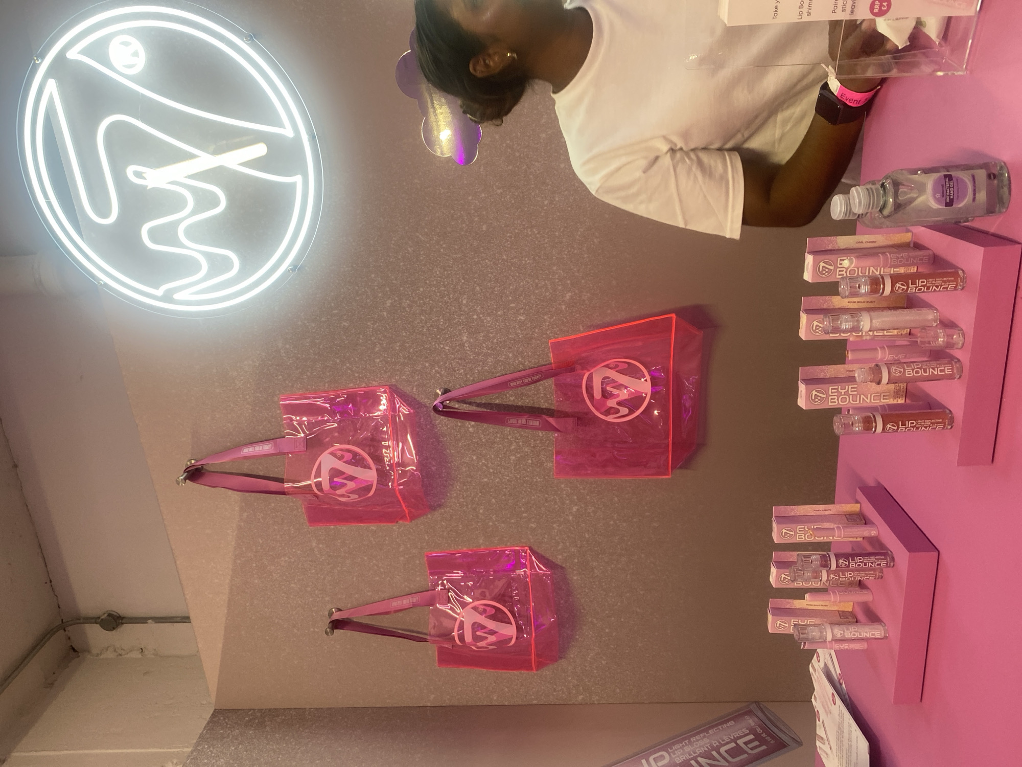 This image captures an indoor scene at a beauty product event or pop-up store in London, United Kingdom. A person with dark skin and hair, wearing a white short-sleeved shirt, a gold earring, and a black smartwatch with a pink "Event" wristband, is visible at the top right, interacting with a display.
The setting features a strong pink aesthetic. Multiple transparent pink tote bags, each bearing a white circular logo, are hung against a speckled light grey wall. The strap of one bag shows the text "HOW WILL YOU BE TODAY?". Below these bags, various cosmetic products are neatly arranged on pink tiered shelves. These products, packaged in tubes and boxes, are branded "BOUNCE", with specific items labeled "LIP BOUNCE" and "EYE BOUNCE". Additional visible text on packaging includes "LIGHT REFLECTING LIP GLOSS" and "BRILLANT À LÈVRES". A clear acrylic stand contains information sheets, with visible phrases like "Take y", "Lip Bo", "shime", "Paire", "stic", "leavi", and a price tag indicating "RRP £4". An "Antibacterial Handgel" bottle is also on the counter.
A prominent white neon sign, displaying a stylized, abstract logo within a circular outline, illuminates the upper left area of the wall. The overall ambiance suggests a modern, vibrant, and organized promotional space, likely for sampling or purchasing beauty items. The presence of the "Event" wristband indicates a specific happening.