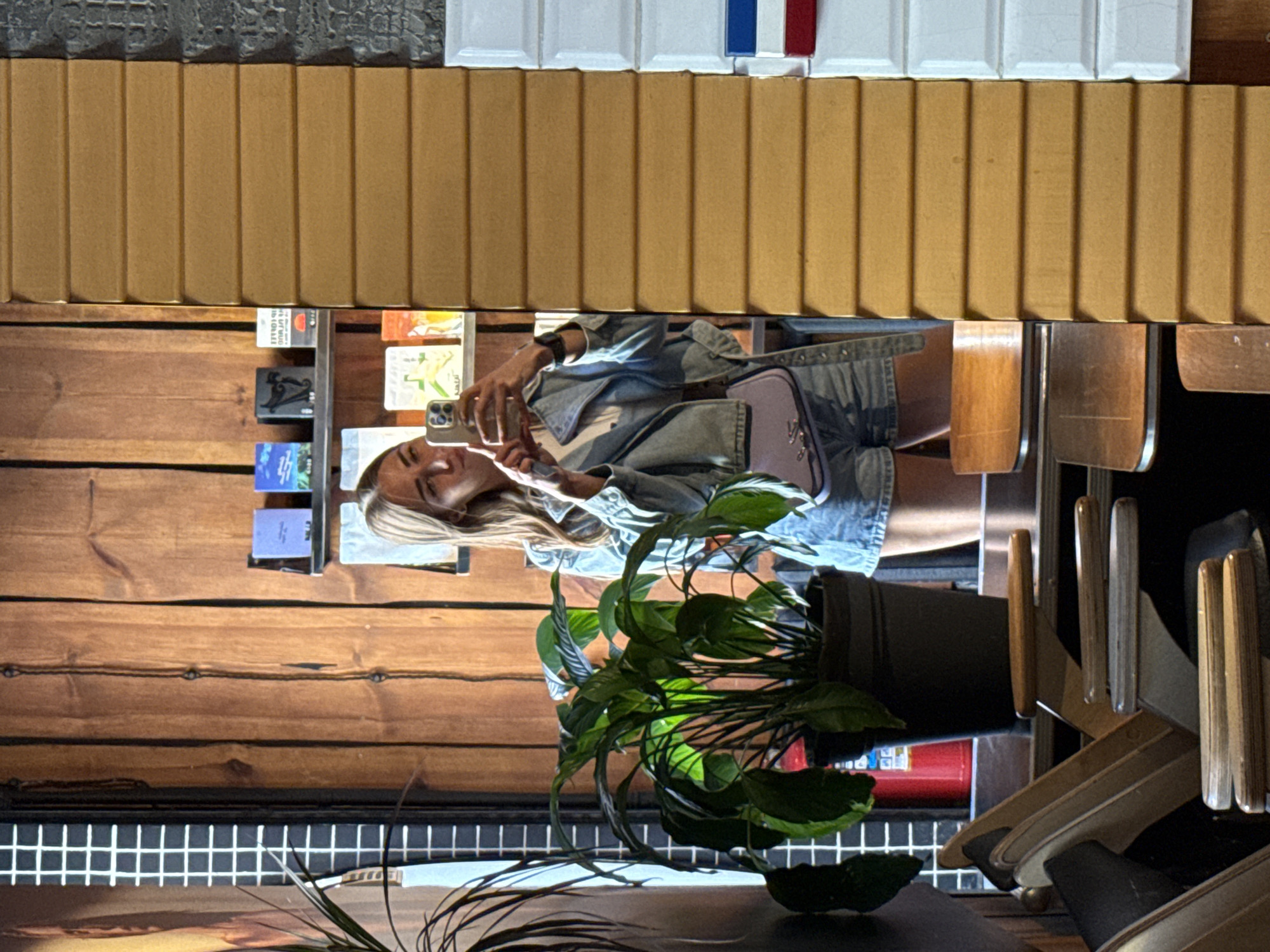 A young woman with blonde hair is taking a selfie in a mirror. She is wearing a denim jacket and shorts, and carrying a small purple purse. She is standing in what appears to be a cafe or restaurant, with wooden walls and shelves displaying various items. In the foreground, there is a large potted plant and several chairs. The overall lighting is warm and inviting. The location is the Cafe Bottega in Saint Petersburg, Russia.