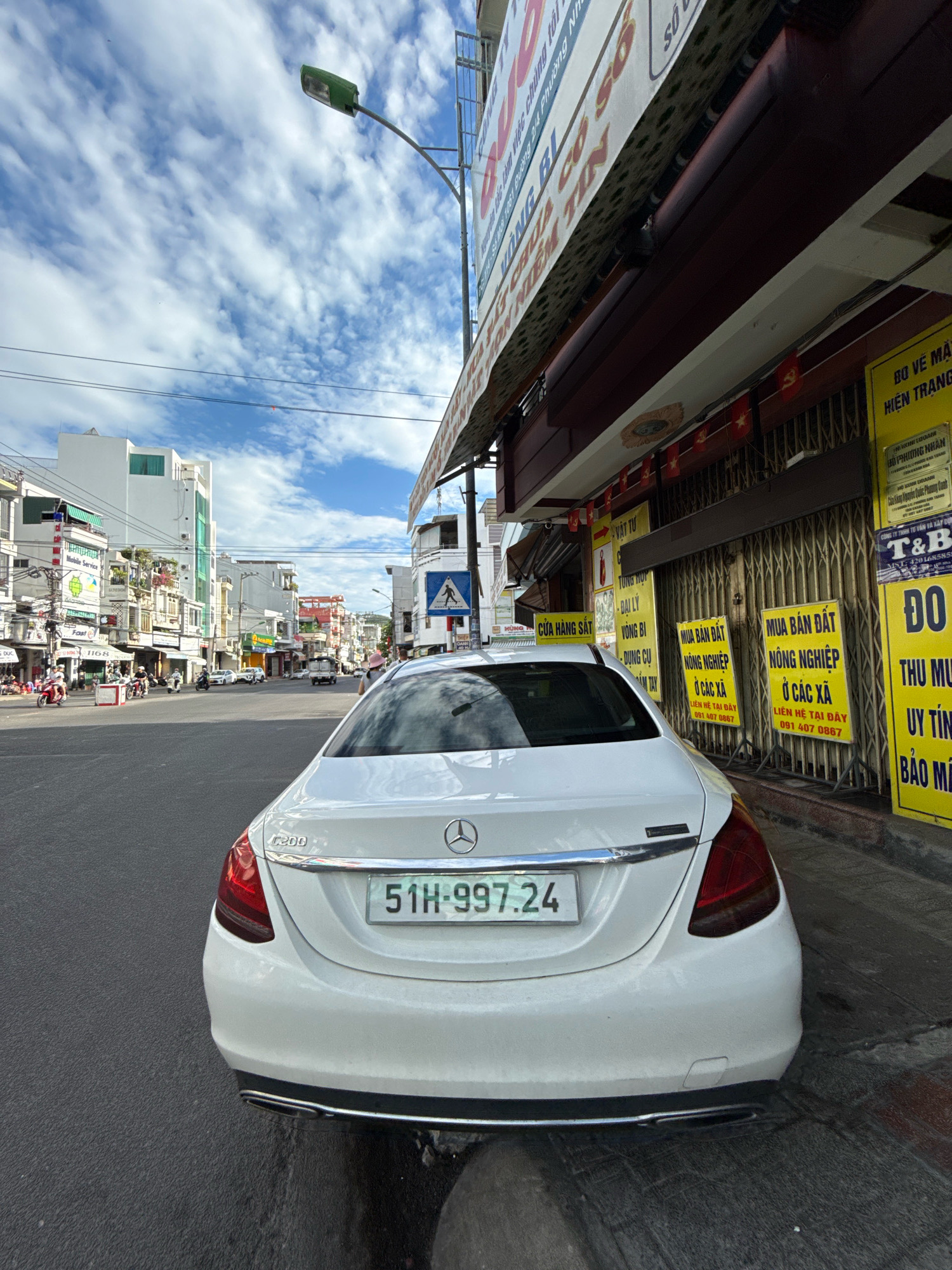 A white Mercedes-Benz C200 sedan with license plate "51H-997.24" is parked on the right side of a street in Nha Trang, Vietnam. The car is viewed from the rear, with its taillights and trunk visible. The street appears to be a commercial area with buildings lining both sides. Several businesses have signs displayed in Vietnamese. On the right side of the street, there are yellow signs advertising "Mua Ban Dat Nong Nghiep O Cac Xa" (Buying Agricultural Land in Villages) with contact phone numbers. The sky is blue with scattered white clouds. A street lamp with a green light fixture is visible in the upper left corner. In the background, there is traffic including motorcycles and cars. The scene suggests a typical urban environment in Vietnam.