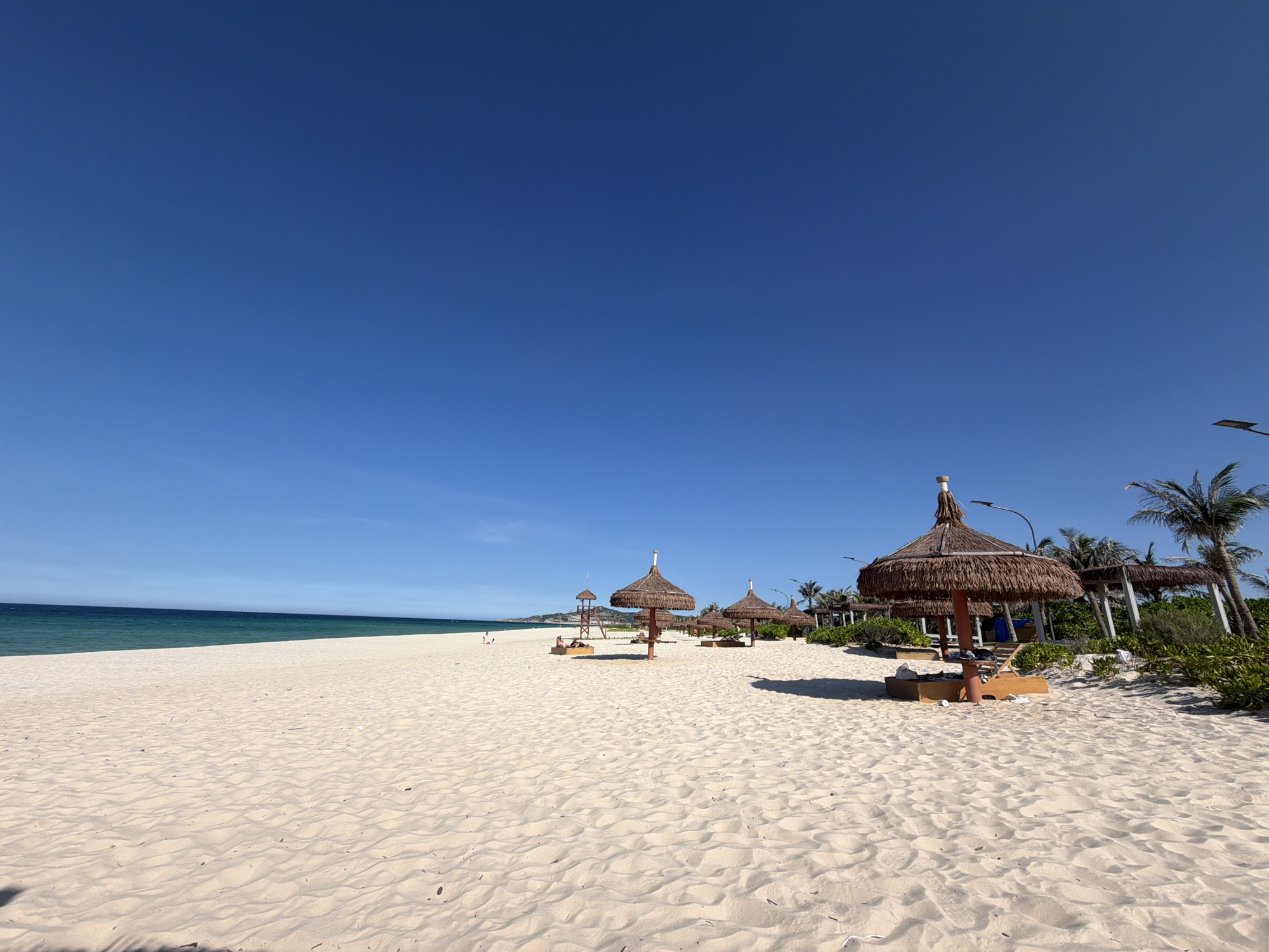 This is a wide-angle, eye-level outdoor photograph of a sandy beach in Cam Ranh Ward, Vietnam. The sky is a clear, bright blue, occupying the upper half of the frame. The beach is composed of light-colored sand with visible ripples and shadows from an unseen light source. In the middle ground, several thatched-roof umbrellas are scattered across the sand, some with lounge chairs underneath. To the left, a lifeguard tower with a red flag is visible. Further into the distance, the turquoise ocean meets the horizon under the blue sky. Palm trees and dense green foliage line the back of the beach. A few scattered people are present on the beach, some near the lifeguard tower and others relaxing under the umbrellas. The scene depicts a tranquil beach resort atmosphere.