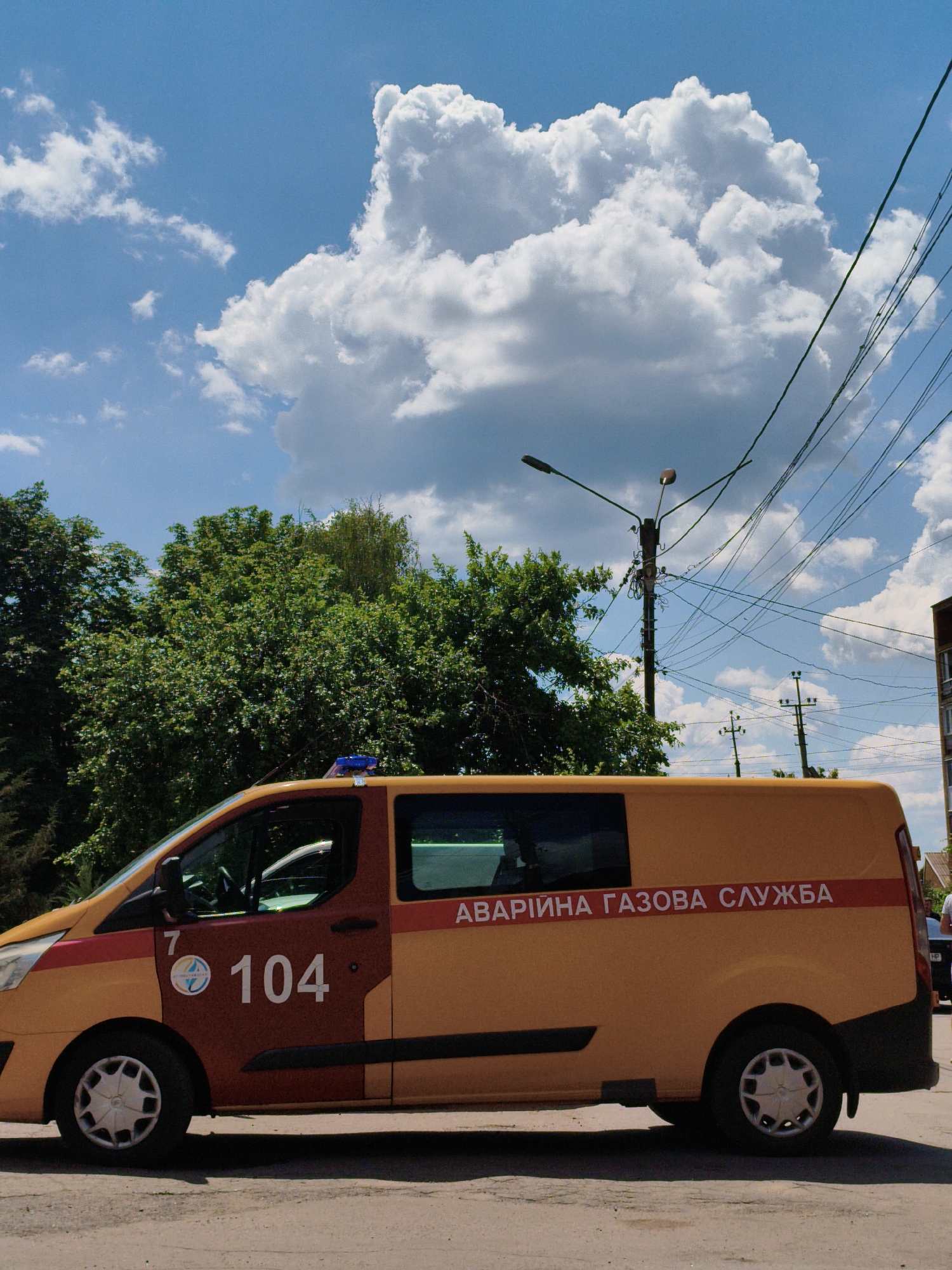 In Velyka Bahachka, Ukraine, a yellow and orange service van with the text "АВАРІЙНА ГАЗОВА СЛУЖБА" (Emergency Gas Service) printed on its side is parked on a street. The van has a blue flashing light on its roof. The side of the van also displays the number "104" and a circular logo. The vehicle is positioned on a paved surface with shadows cast by the sun. Behind the van, a line of green trees is visible under a bright blue sky with large, white cumulus clouds. Utility poles with a complex network of wires are also prominent features of the environment. The scene appears to be set during daylight hours.