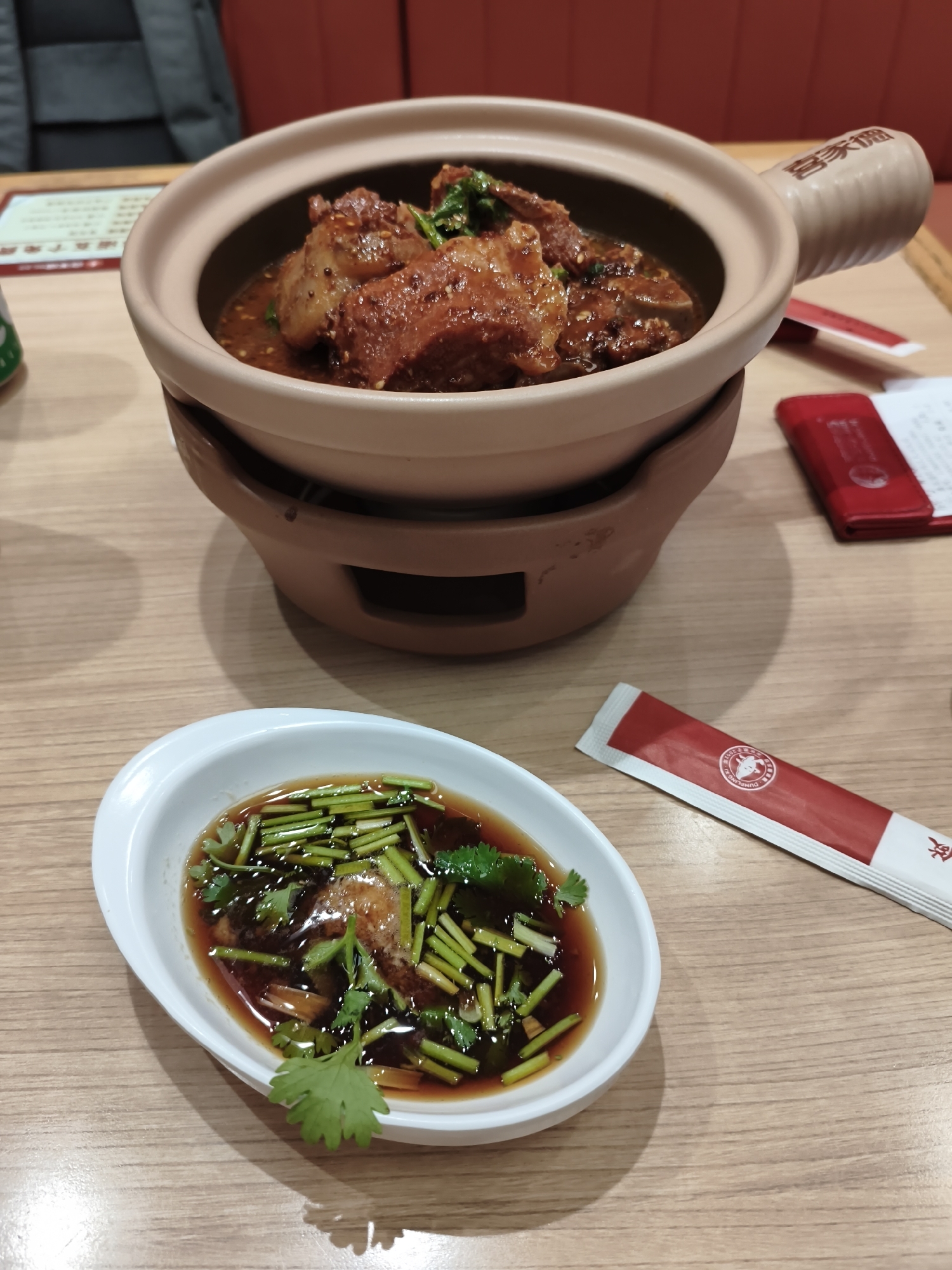 The image shows a close-up view of a meal served at a restaurant. In the foreground, there is a small white dish containing a dark sauce garnished with chopped green onions and cilantro. Behind this dish, a larger clay pot with a light brown exterior and a darker interior holds a stewed meat dish. The pot has two handles and sits on a brown stand. To the right of the clay pot, a red packet, possibly containing chopsticks or utensils, lies on the wooden table. A red card or menu is also visible further to the right. The background shows a blurred out red booth seating area and a table. The overall setting suggests a casual dining environment. The image does not contain any identifiable information or features that would allow for pinpointing its location to the city of Mudanjiang, China.
