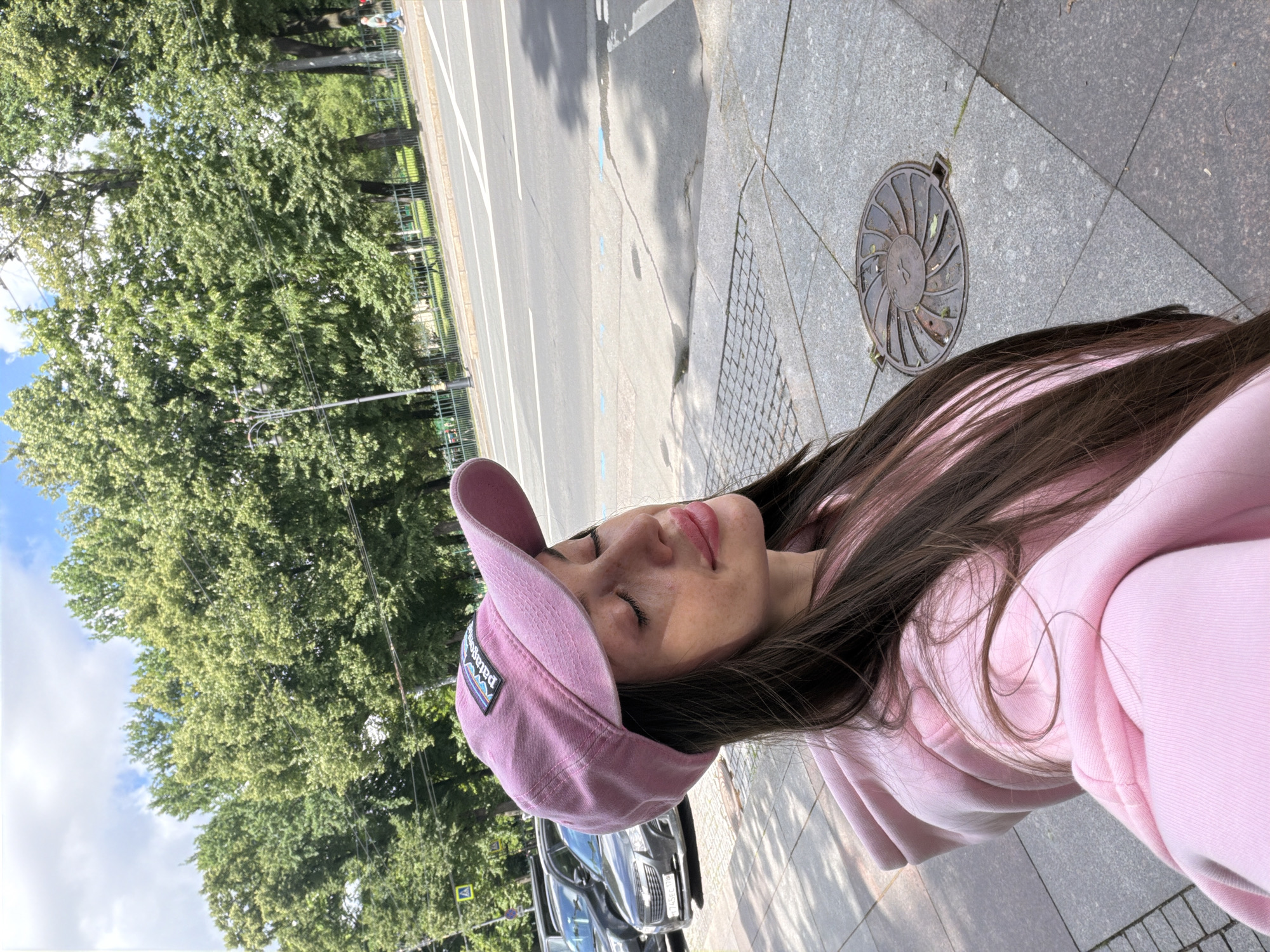 A young woman with long, dark hair and some pink highlights stands on a sidewalk in Saint Petersburg, Russia, with her eyes closed and head tilted back. She is wearing a pink baseball cap with a Patagonia logo and a pink hoodie. The background shows a street with cars, trees, and a park.