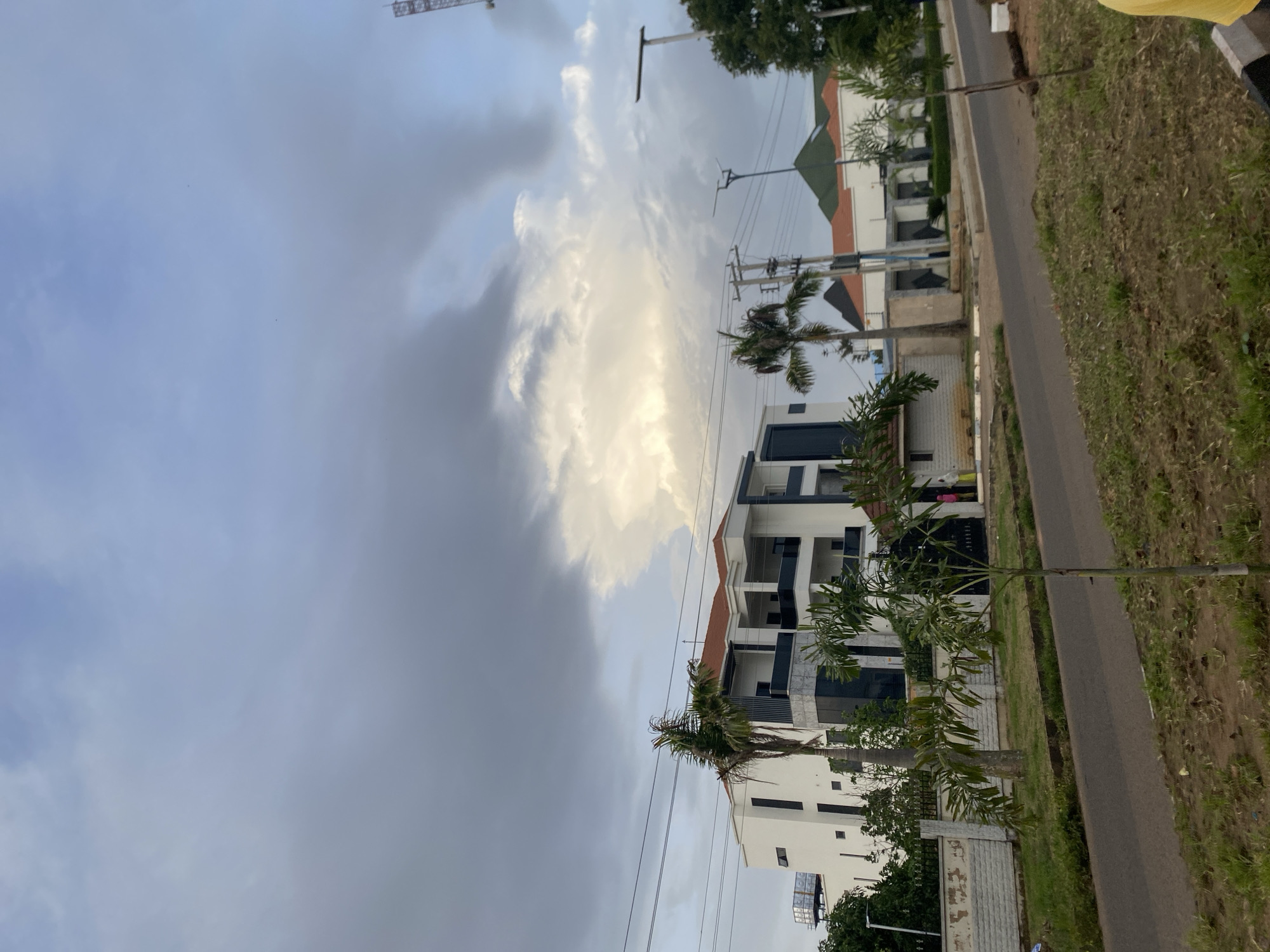 This image captures a scene in Barikin Jirga Summa, Nigeria, on a day with dramatic cloud formations dominating the sky. In the foreground, a paved road runs horizontally across the frame, bordered by grassy verges with scattered dry patches and young trees. On the left side of the road, a modern, multi-story white building with dark window accents and orange roof trim stands prominently, partially obscured by a tall palm tree. A low, textured white wall with a black metal fence separates the building from the road. Further down the road, another building with a green roof and orange trim is visible. The sky is filled with a mix of dark grey and bright white clouds, suggesting changing weather conditions. Power lines and utility poles are visible, crossing the sky above the buildings. Two individuals, one wearing pink and the other yellow, are standing near the entrance of the building on the left. The overall impression is of a residential or commercial area under an overcast sky.