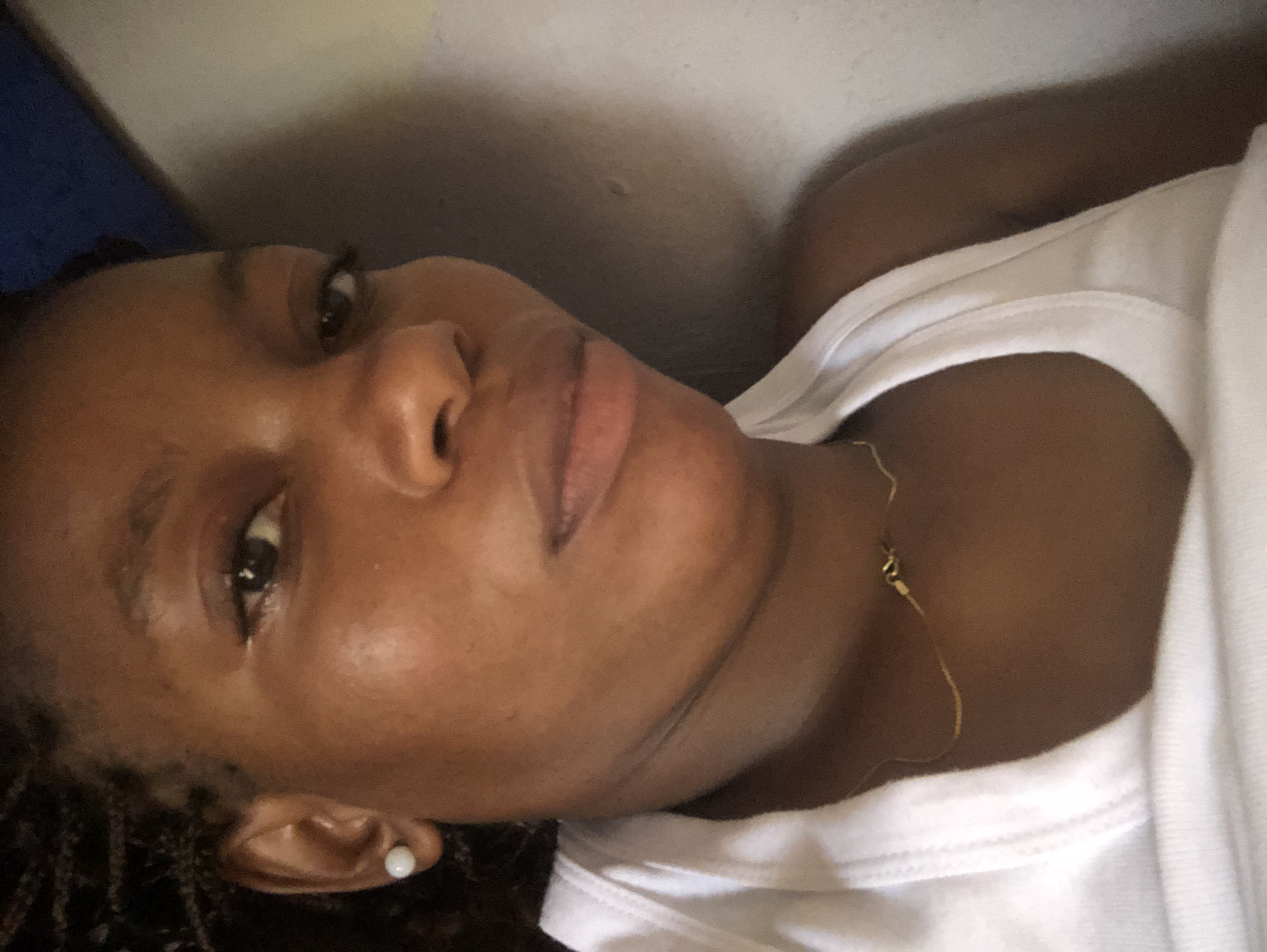 A close-up, indoor photograph shows a Black woman with dark brown skin and braided hair. She is wearing a white top and a gold necklace. A white stud earring is visible in her left ear. She appears to be lying down. The background consists of a pale wall, and a dark blue object is visible in the upper left corner. There are no indications as to the location of the photograph being Kashere, Nigeria.