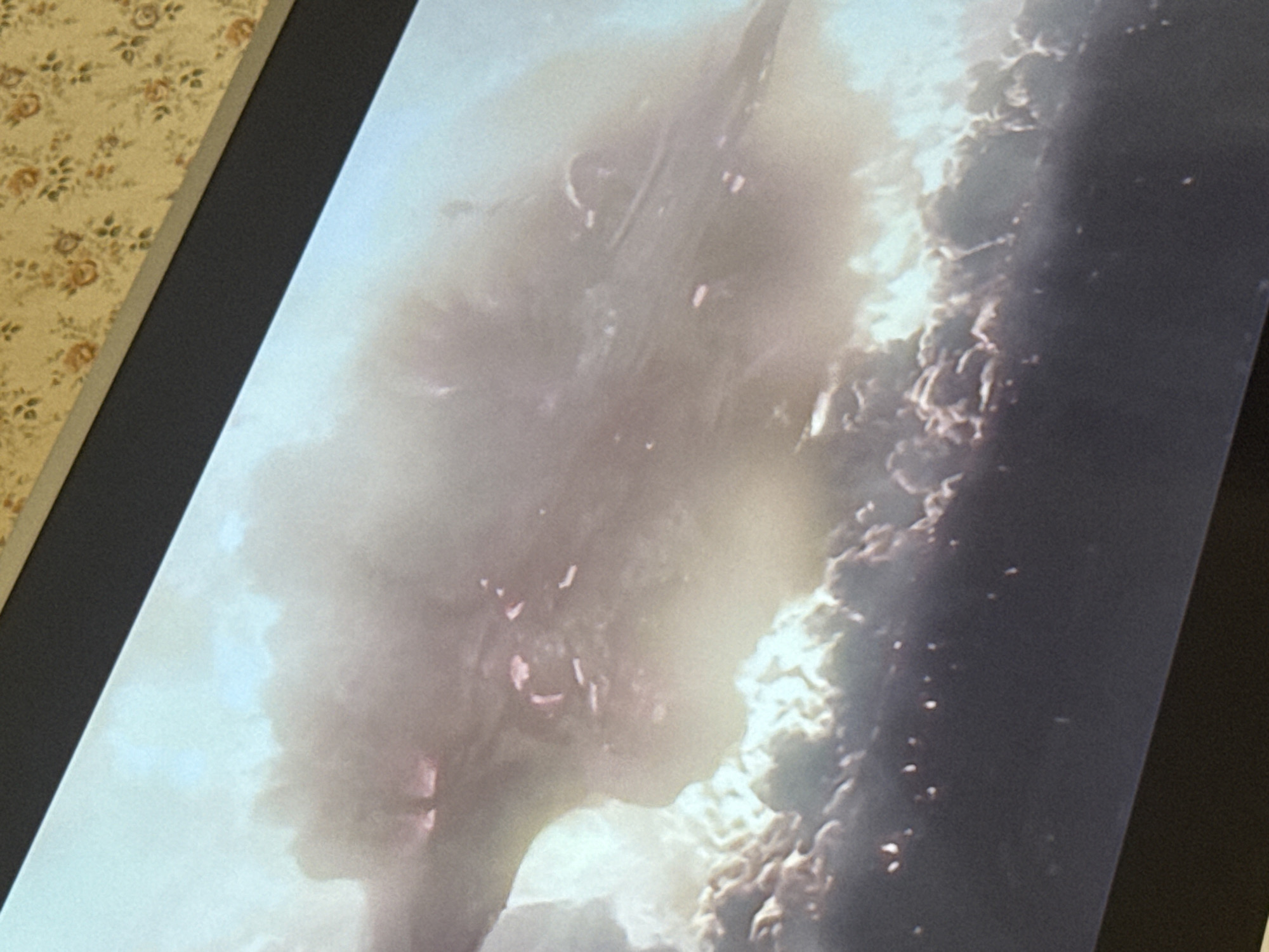 The image shows a section of a screen displaying an indeterminate scene. On the left, a diffused, bright area obscures details. To the right of this area, there are suggestions of texture and possibly objects. The background is dark, and the image is viewed at an angle, with a patterned wall visible to the left of the screen. No people, actions, or interactions are evident. There is no indication of the city of Mytishchi, Russia.