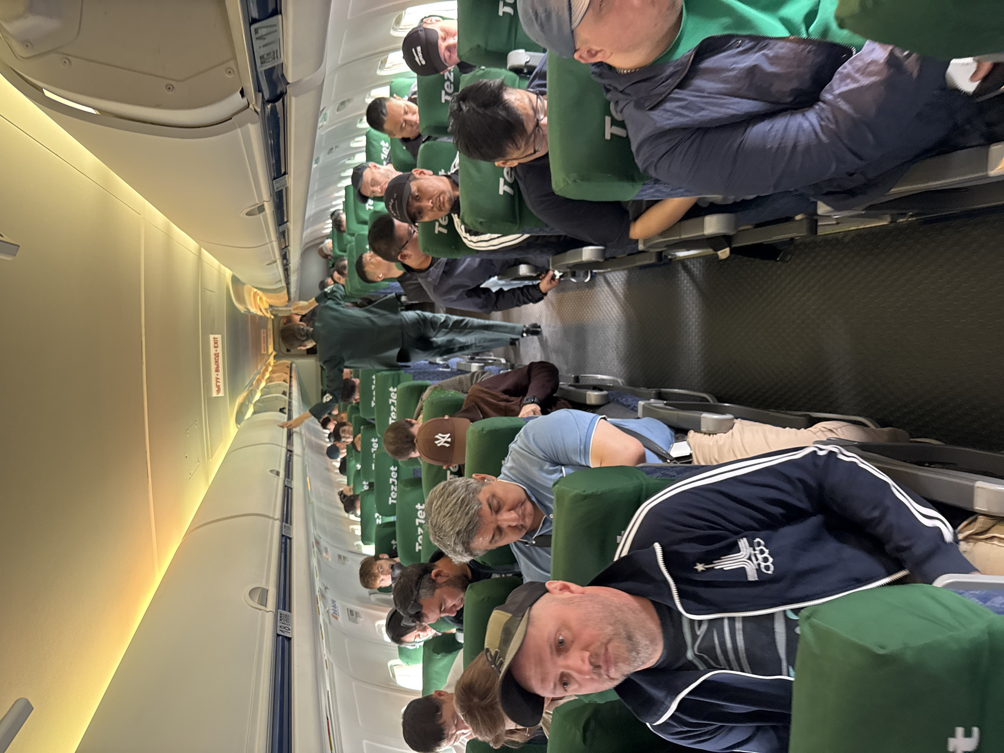 The image shows the interior of an airplane cabin. Passengers are seated in rows, most with their heads resting against the headrests, likely asleep or relaxing. The seats are green and have the word "TezJet" printed on them. Overhead compartments line the top of the cabin. An emergency exit sign, with text in Russian and English, is visible near the aisle. The overall environment suggests a packed flight, possibly during a long-distance travel. There is no explicit information about Tashkent, Uzbekistan in the image.