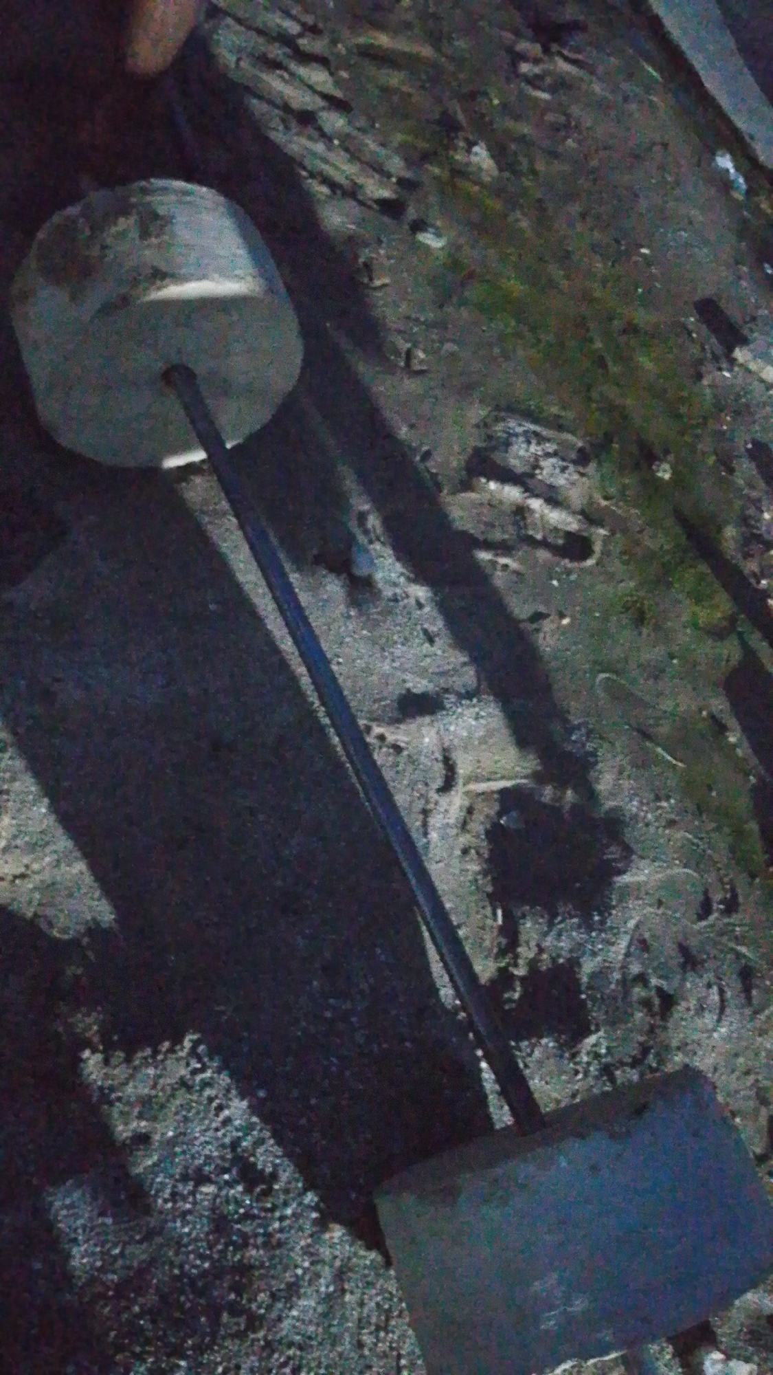 The image shows a makeshift barbell constructed from concrete weights and a metal bar, lying on a rough, possibly dirt or gravel surface. The scene appears to be outdoors, with elements of a distressed or unkempt environment, possibly a yard or an unused area. One concrete weight, circular in shape, is visible on the left, with the metal bar extending diagonally across the frame. Another concrete weight, rectangular in shape, is visible at the bottom right, with the bar inserted into it. The lighting suggests it might be dusk or night, casting long shadows. The background appears to be a mix of dirt, rubble, and sparse vegetation. There is no indication of the city of Indo, Nigeria within the image itself.