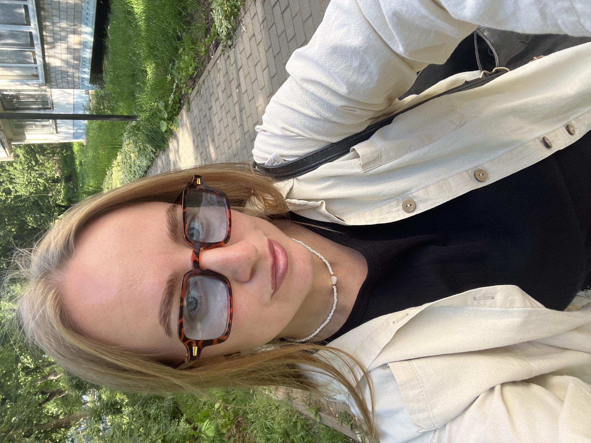 This is a close-up, head-and-shoulders selfie of a young woman with blonde hair, wearing tortoiseshell glasses, a black top, and a light-colored open shirt. She is also wearing a delicate beaded necklace. The photo appears to be taken outdoors on a sunny day, indicated by the bright lighting and shadows. The background shows a paved walkway, green grass, and foliage, suggesting a park or residential area. In the distance, a building with windows is visible. The woman's expression is neutral. The image is oriented sideways.