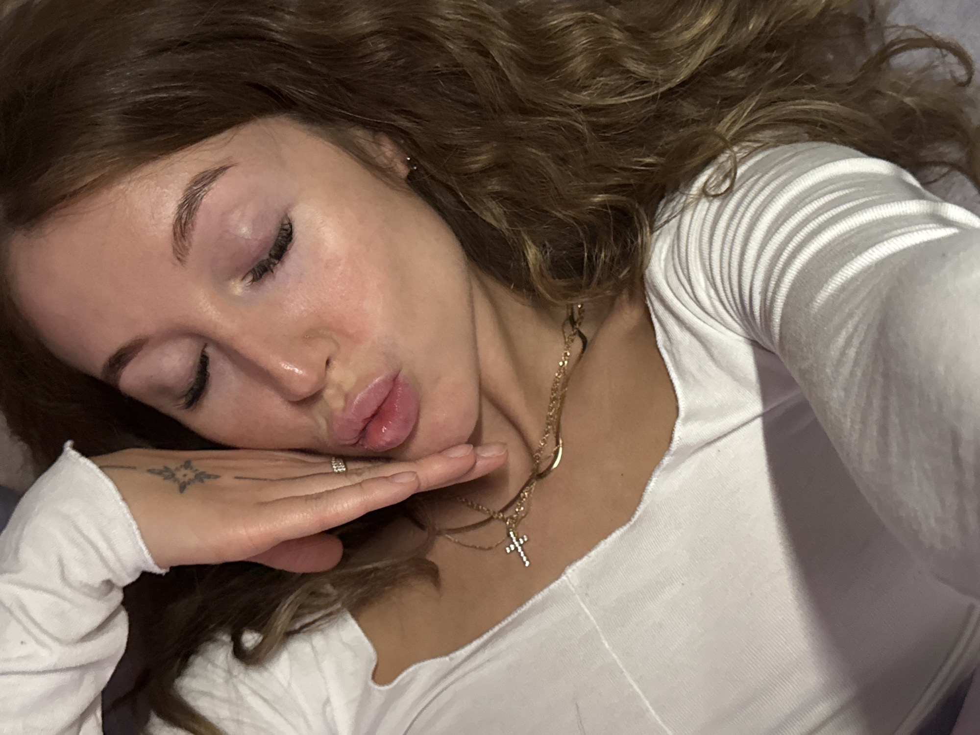 A young woman is lying down, presumably indoors, wearing a white long-sleeved shirt and a layered gold necklace with a cross pendant. Her eyes are closed, and she is making a kissing expression with her lips. Her hand is positioned under her chin. There is a tattoo on the back of her hand. Her curly hair is spread out around her head. This image does not provide information about the location being in the city of Chaikovsky, Russia. The description focuses on the observable elements in the image.