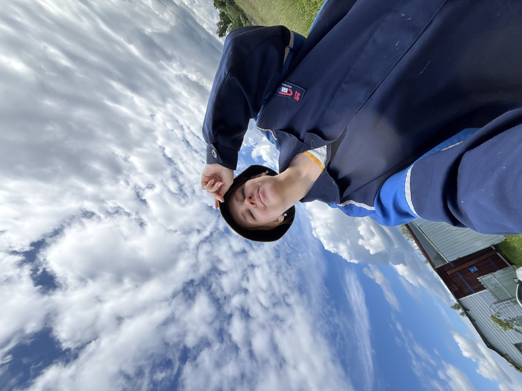 The image shows a young woman taking a selfie outdoors. She is wearing a dark blue jacket with blue and white stripes on the sleeves, and a dark bucket hat. The woman is looking towards the camera with a slight smile and her right hand is touching the brim of her hat. The background features a bright blue sky filled with white, fluffy clouds. To the left of the woman, there is a rustic-looking building with a corrugated metal exterior and a dark-colored door. The ground appears to be covered in grass, and there are trees visible in the distance to the right. The scene suggests an outdoor setting in a rural or semi-rural area, potentially in the city of Дунилово, Russia, given the context provided. The overall lighting indicates it is daytime.