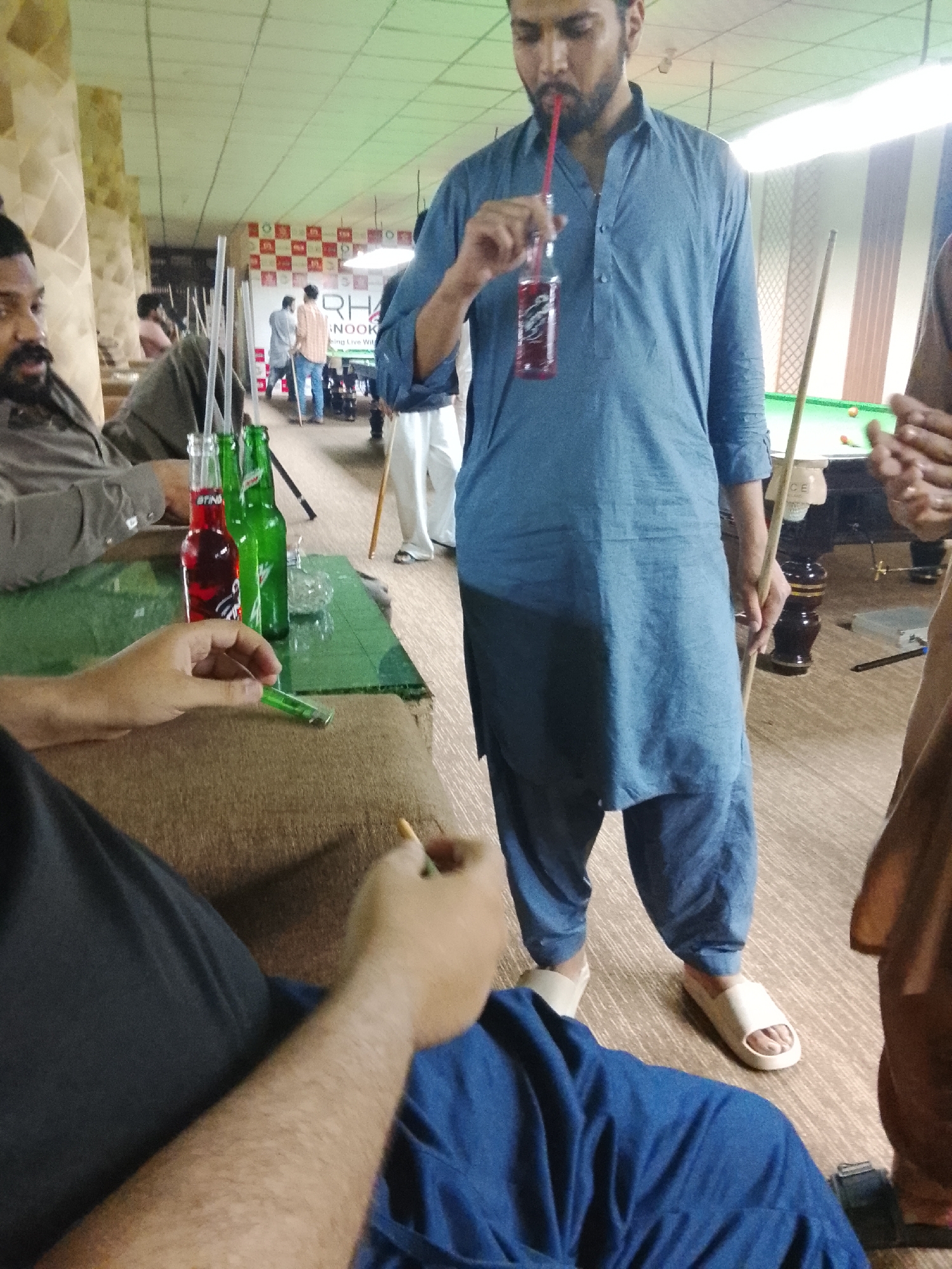 Inside a pool hall in Unknown, Unknown, a man in a blue tunic and white slippers stands, drinking from a glass bottle with a red straw while holding a pool cue. Another man, seated on a cushioned bench, holds a cigarette in one hand and a lighter in the other. On the green glass table between them are bottles of soda, one red and two green, along with straws and a clear ashtray. In the background, other patrons are seen playing pool. The room is illuminated with fluorescent lighting.