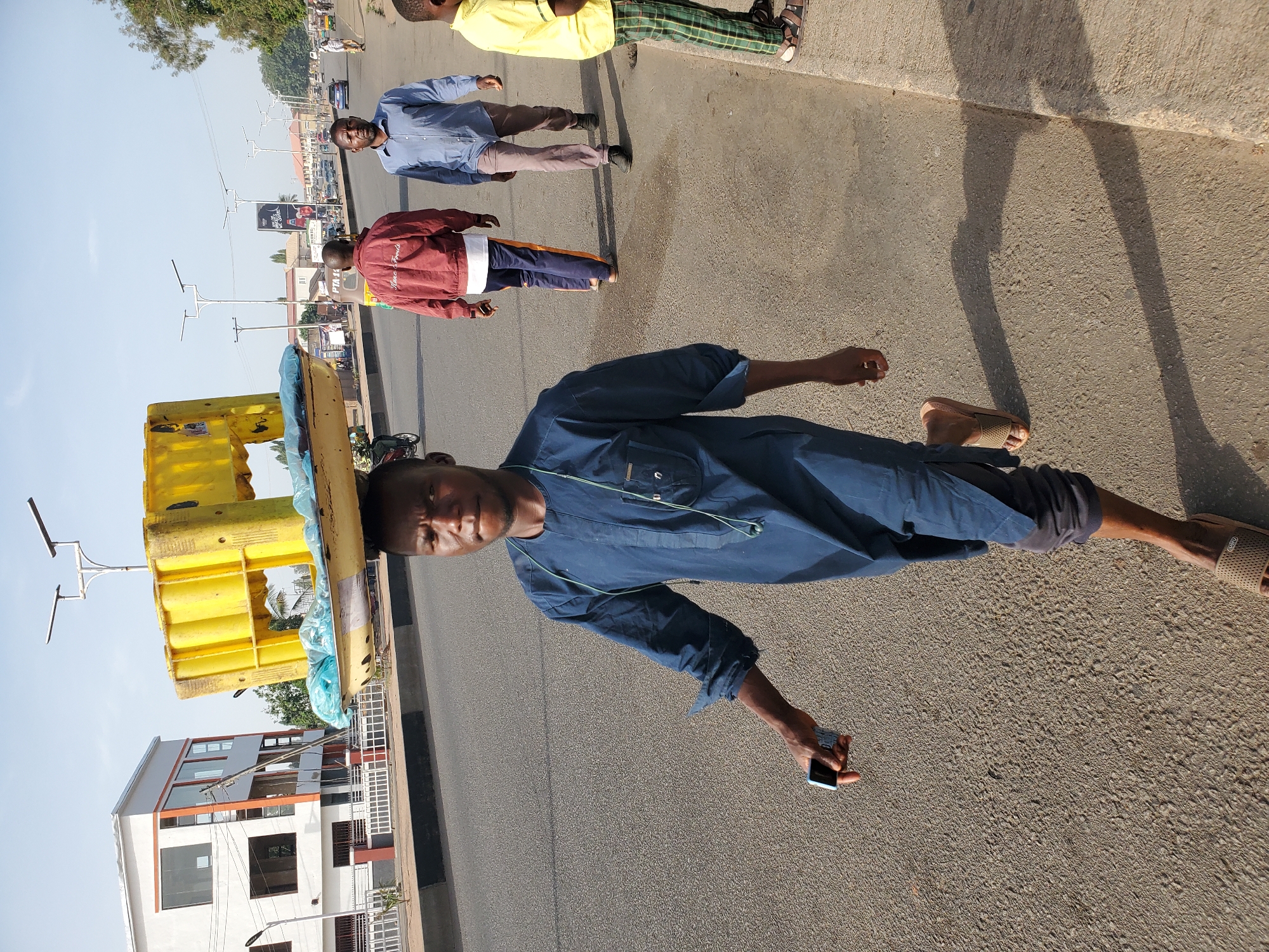 This media file captures a vibrant street scene in Jos, Nigeria, under bright daylight. The main subject in the foreground is a man wearing a dark blue long-sleeved tunic and dark trousers, with a light brown sandal on his visible foot. He carries a large, bright yellow, box-like structure, possibly a cooler or vendor's display unit, balanced on his head, with some blue fabric or plastic visible around it. He holds a black smartphone in his right hand and looks directly at the camera.
Several other individuals are visible on the paved street and sidewalk in the mid-ground. To the right, a man with a beard, wearing a light blue button-down shirt and light brown trousers, stands on the sidewalk. Behind him, another man in a red jacket with the words "True Friends" clearly visible on the back, and dark trousers with side stripes, is also on the sidewalk. Further back and to the right, a man in a yellow shirt and green plaid trousers can be seen.
The setting includes a wide, paved road with a concrete sidewalk. Streetlights with horizontal arms line the street, stretching into the distance. In the background, various buildings are visible, including a prominent white building with orange trim and multiple windows on the left. A large billboard or sign is partially visible further down the street on the right, with "PTA" faintly legible. Trees and other foliage are seen in the distance, suggesting a somewhat green urban environment.
The strong, distinct shadows cast by the people and objects indicate a sunny day, likely late morning or early afternoon. The overall activity depicts people going about their daily routines in a bustling street environment, with the foreground individual appearing to be a mobile vendor.