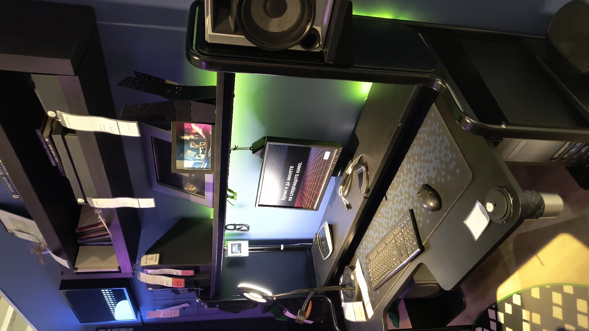 The image depicts a computer setup, seemingly part of a product display or exhibition. It includes a desk with a mouse, keyboard, and monitor, complemented by shelving filled with books and other display items. The setup is accented with green and blue lighting. A closer look at the monitor shows text written in Bulgarian, including phrases like "Време е да минете на следващото ниво," which translates to "It's time to move to the next level." The scene suggests a modern or technologically focused display. Given the Bulgarian language on the monitor, it's likely the location is in Bulgaria, specifically Sofia.